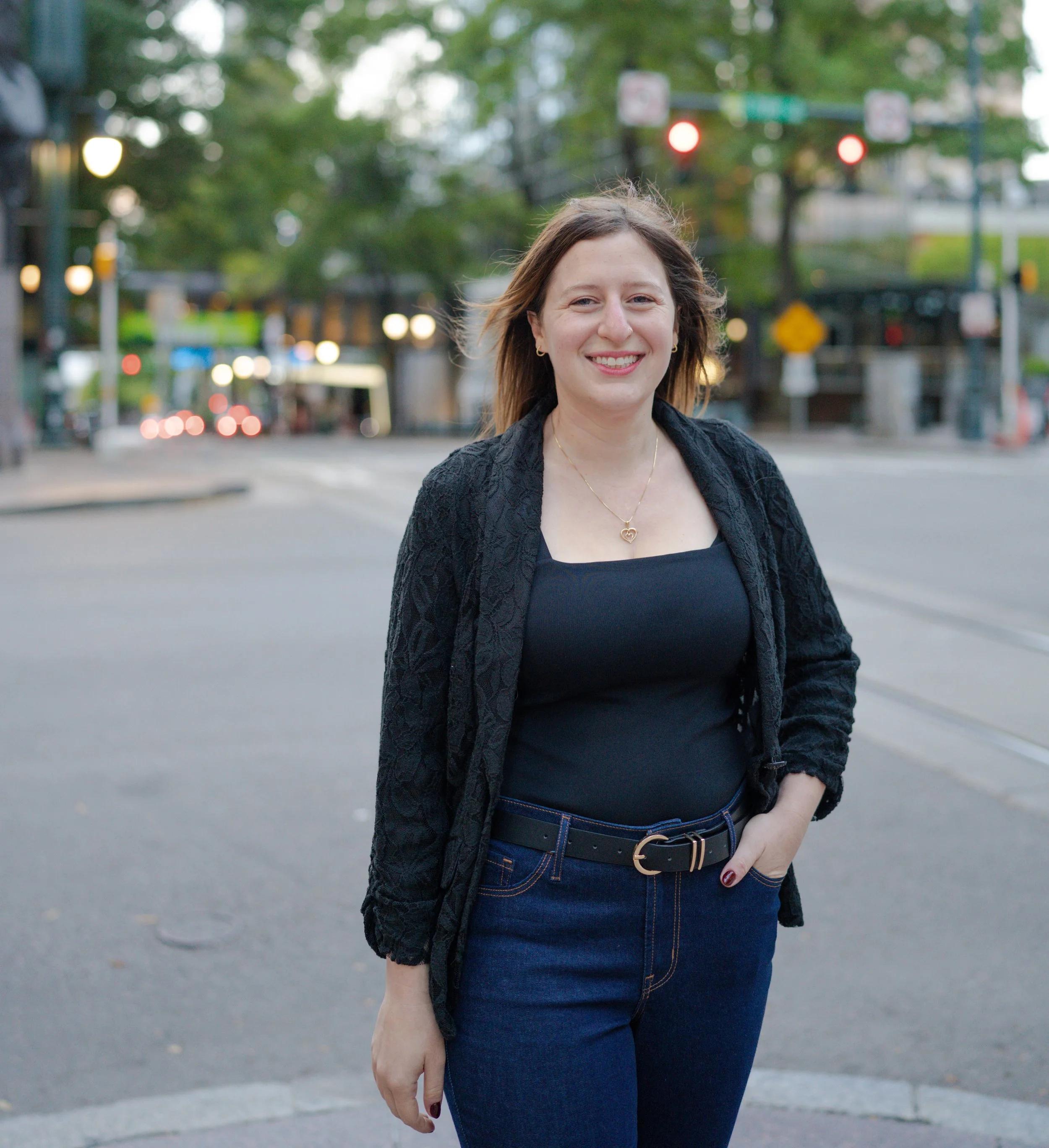 A smiling woman with shoulder-length brown hair, wearing a black top, a black lace jacket, and blue jeans, standing on a city street with traffic lights and trees in the background.