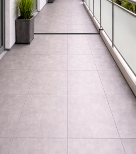 Balkon mit großen grauen Fliesen, Pflanzen in Pflanzkübeln und Glasgeländer.