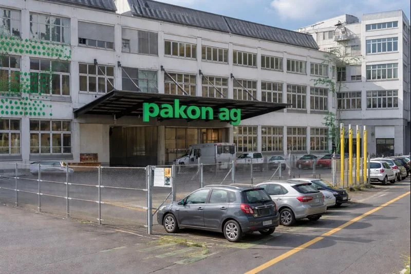 Bürogebäude mit einem Eingang, der von einem Überhang beschattet wird, und einem Parkplatz mit mehreren Autos.