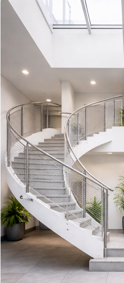 Innenaufnahme eines modernen Gebäudes mit einer kreisförmigen, weißen Treppe mit Metallgeländer und Pflanzen im Vordergrund.