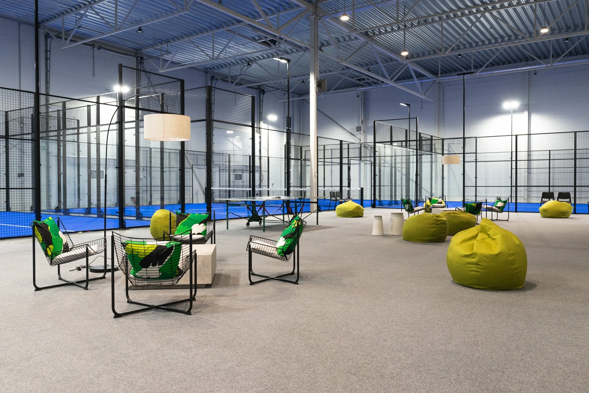 An indoor sports facility with enclosed courts for padel, featuring black fencing, blue courts, and seating area with chairs, bean bag chairs, and colorful cushions.