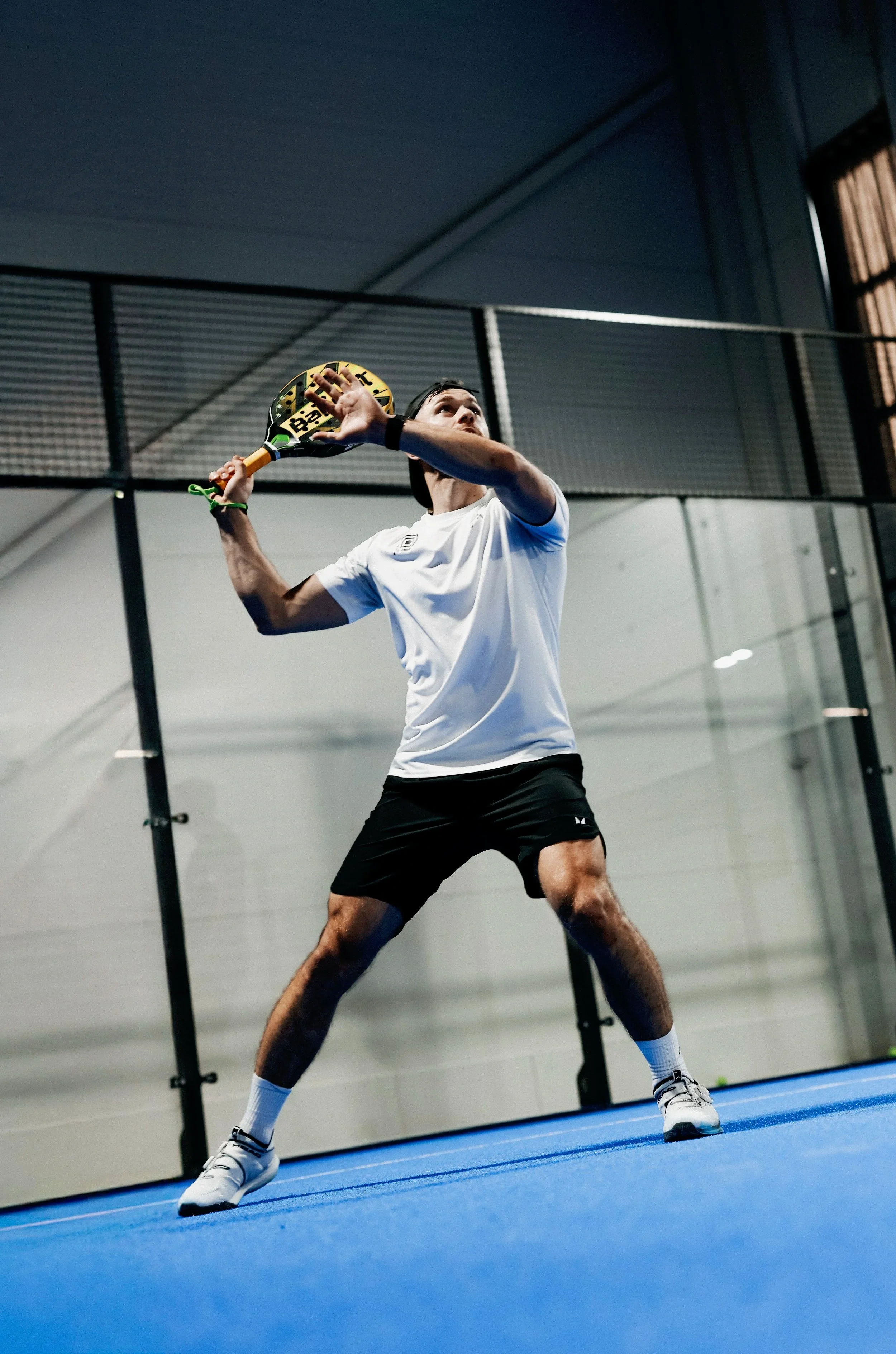 Padel player in white shirt, black shorts, and white shoes preparing to hit a ball with a yellow racket on a blue indoor court.