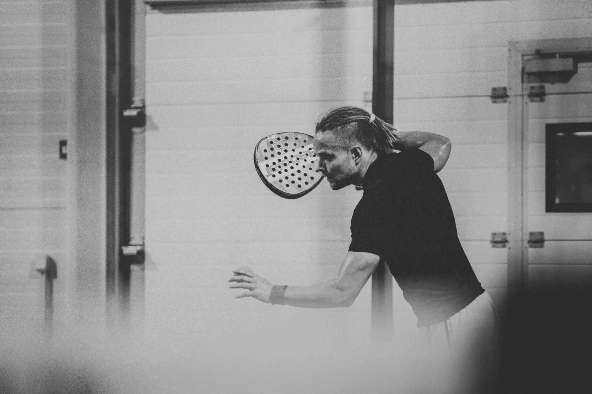 A man in black sportswear preparing to hit a padel ball with a racket in an indoor sports facility.