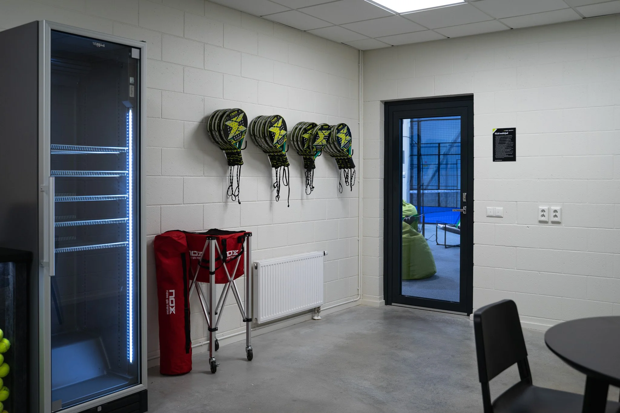 Indoor sports locker room with a glass door leading outside, wall-mounted tennis racket grips, a red carrying case, a folding hand truck, a blue sports drink fridge, and a round black table with black chairs.