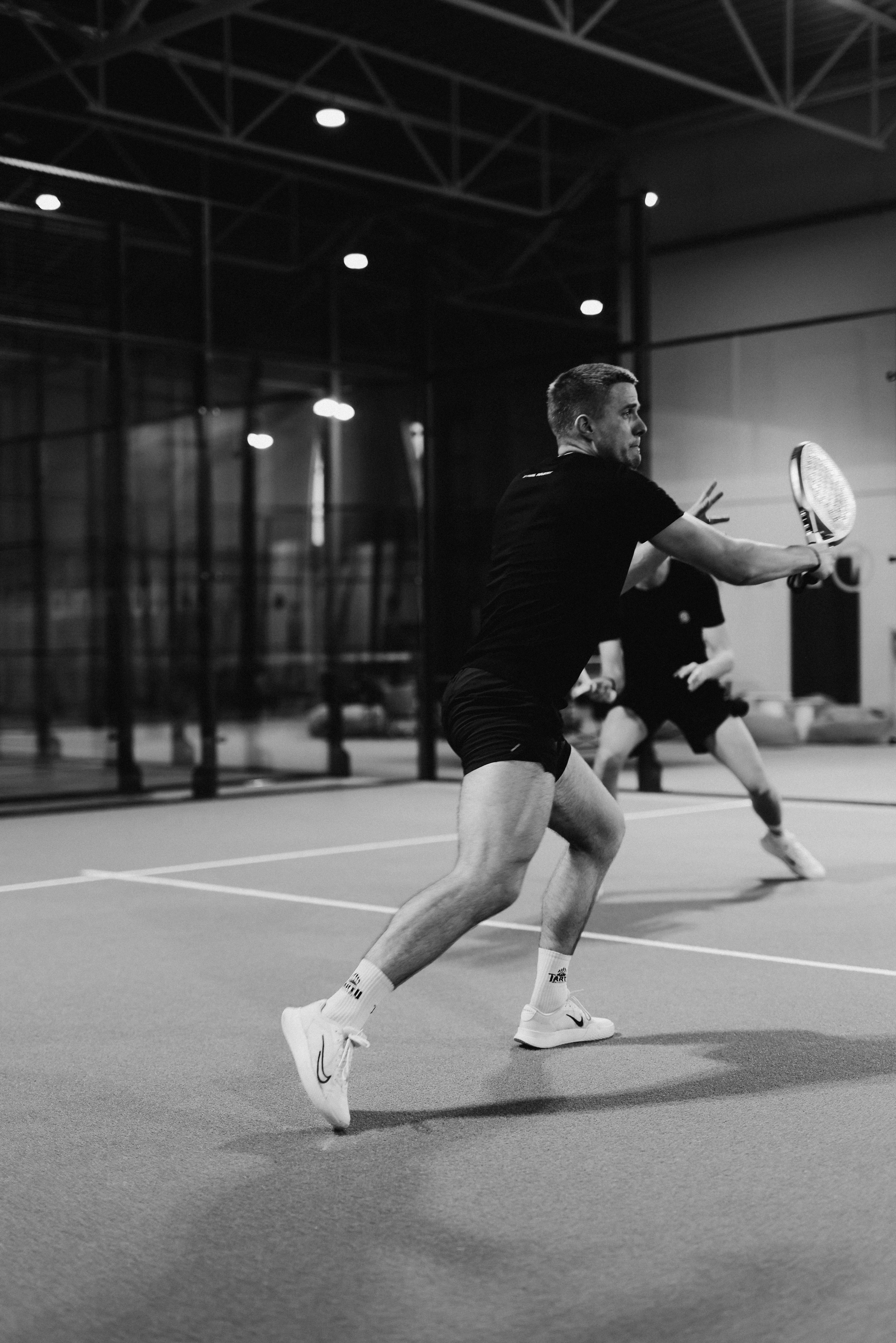 A man  playing padel indoors on a court with a glass wall in the background; the man is in the foreground preparing to hit a ball with a racket.