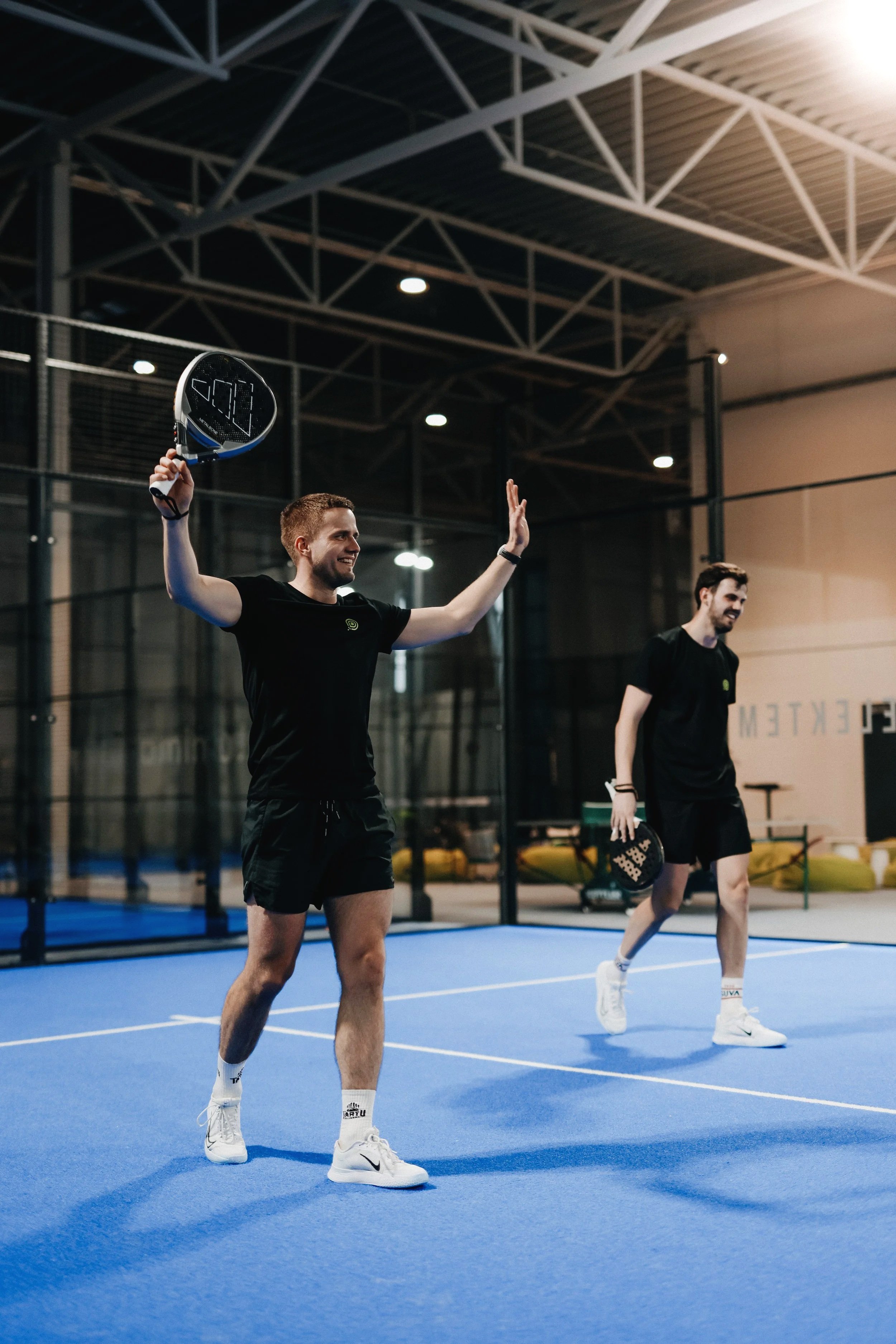 Two men playing pickleball on an indoor court, one holding a racket and celebrating, the other walking with a paddle, both dressed in black athletic wear.
