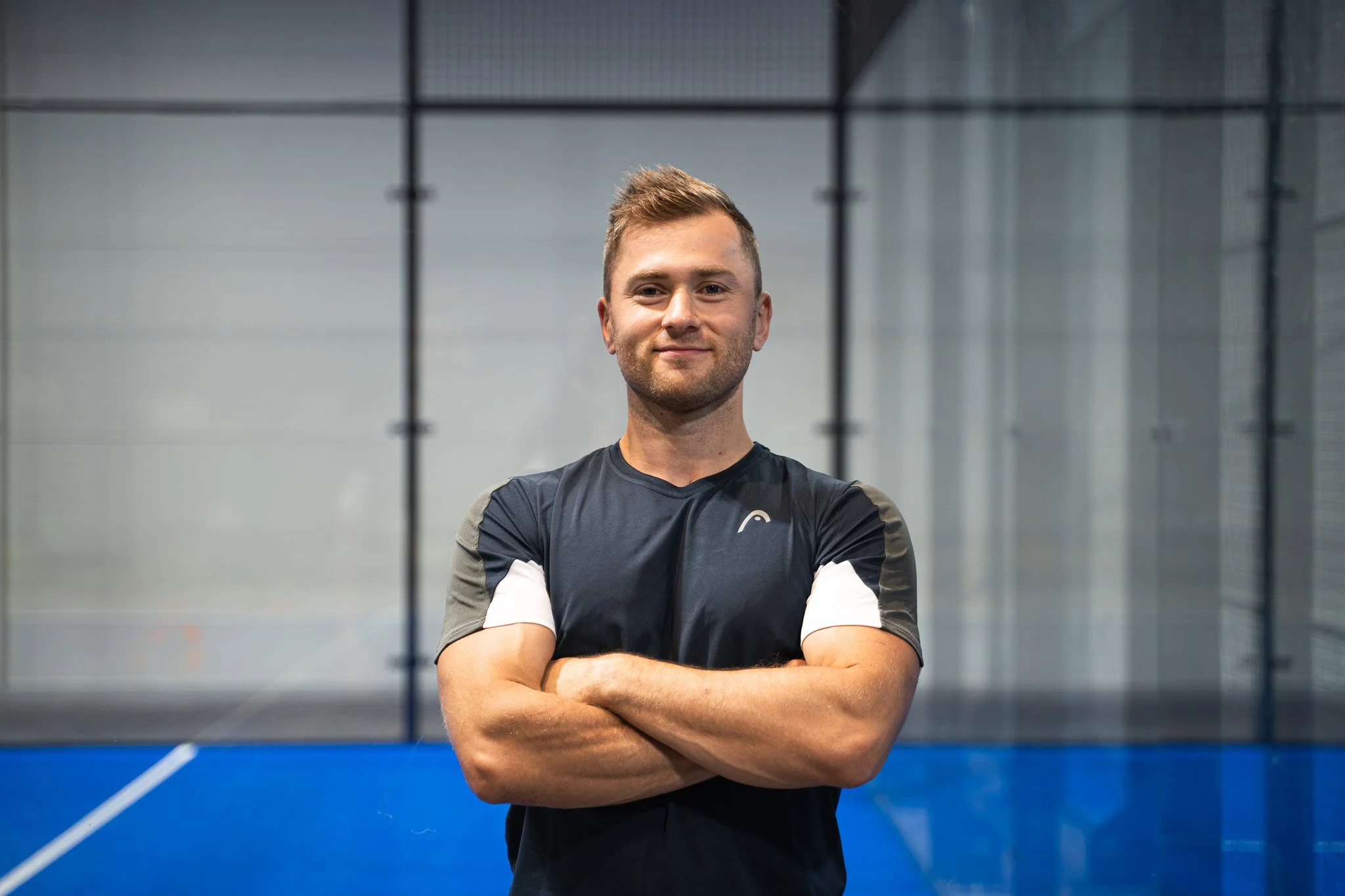 A young man in sports attire standing with arms crossed inside an indoor sports facility with a blue court and glass walls.