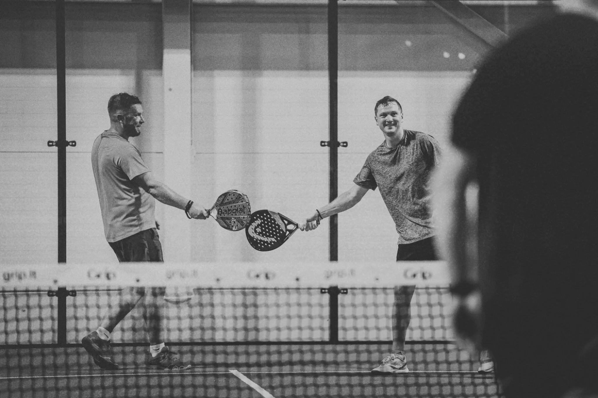 Two men playing padel on an indoor court, shaking hands over the net, with a blurred player in the foreground.