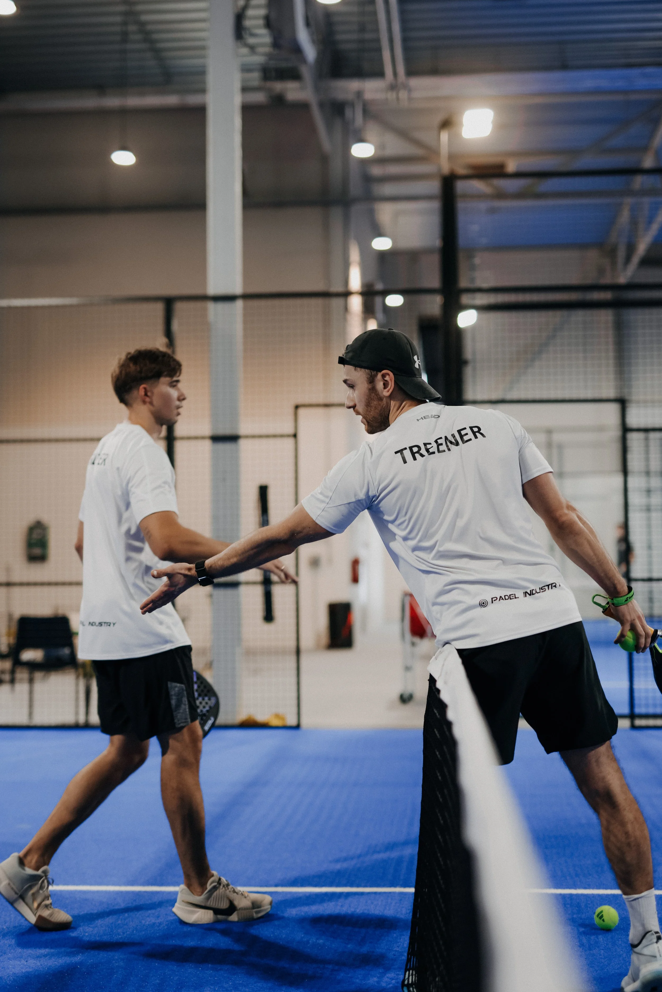 A padel coach in a white shirt with 'Trainer' written on the back coaching a young player on a blue padel court indoor.
