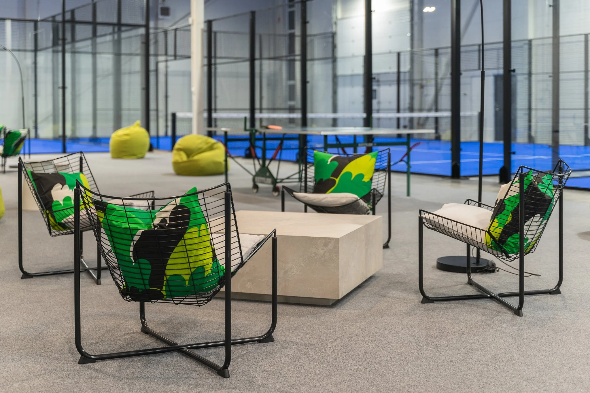 Indoor lounge area with black wire chairs, colorful cushions with green, black, and white abstract designs, a rectangular stone table, and bean bags in front of glass-walled padel courts.