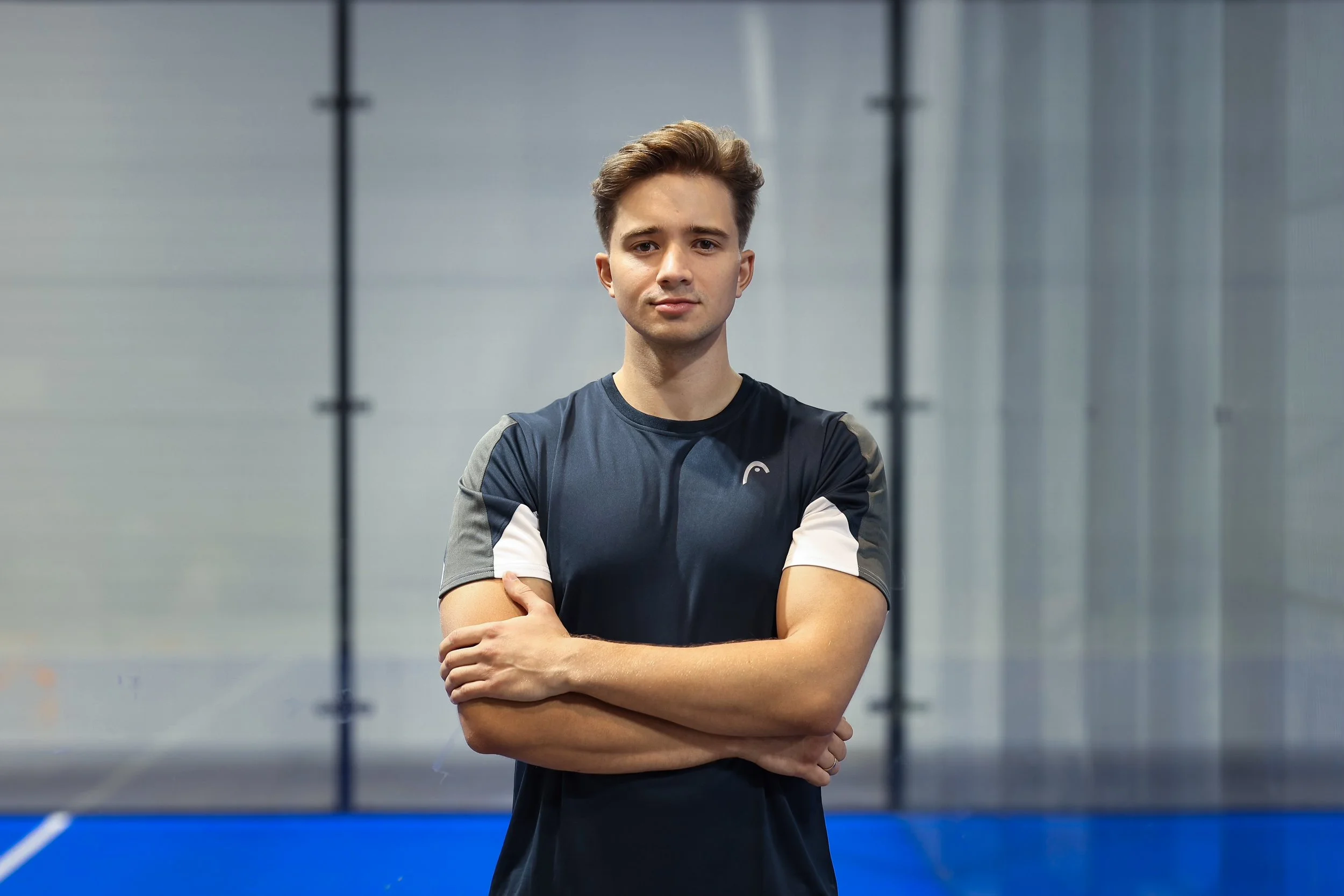 Young man in athletic wear standing with arms crossed in an indoor sports facility.