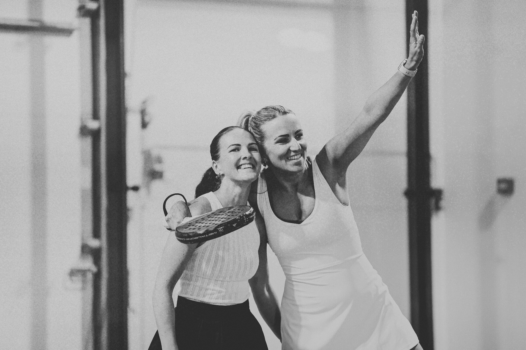 Two women smiling and celebrating after playing padel in an indoor sports facility. One woman is holding a racket.