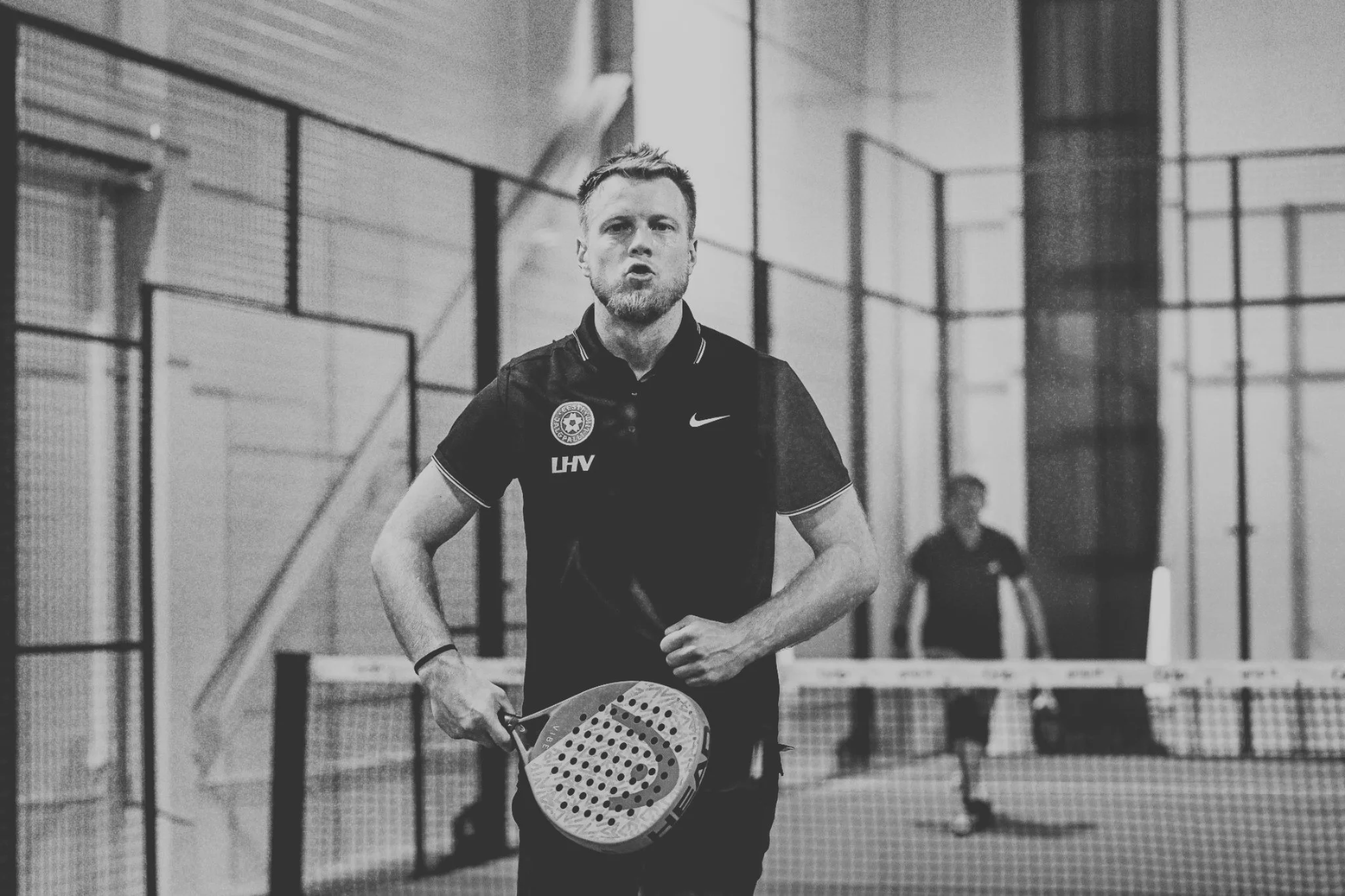 A man playing padel on an indoor court, holding a racket, with a net in front of him and another player in the background.