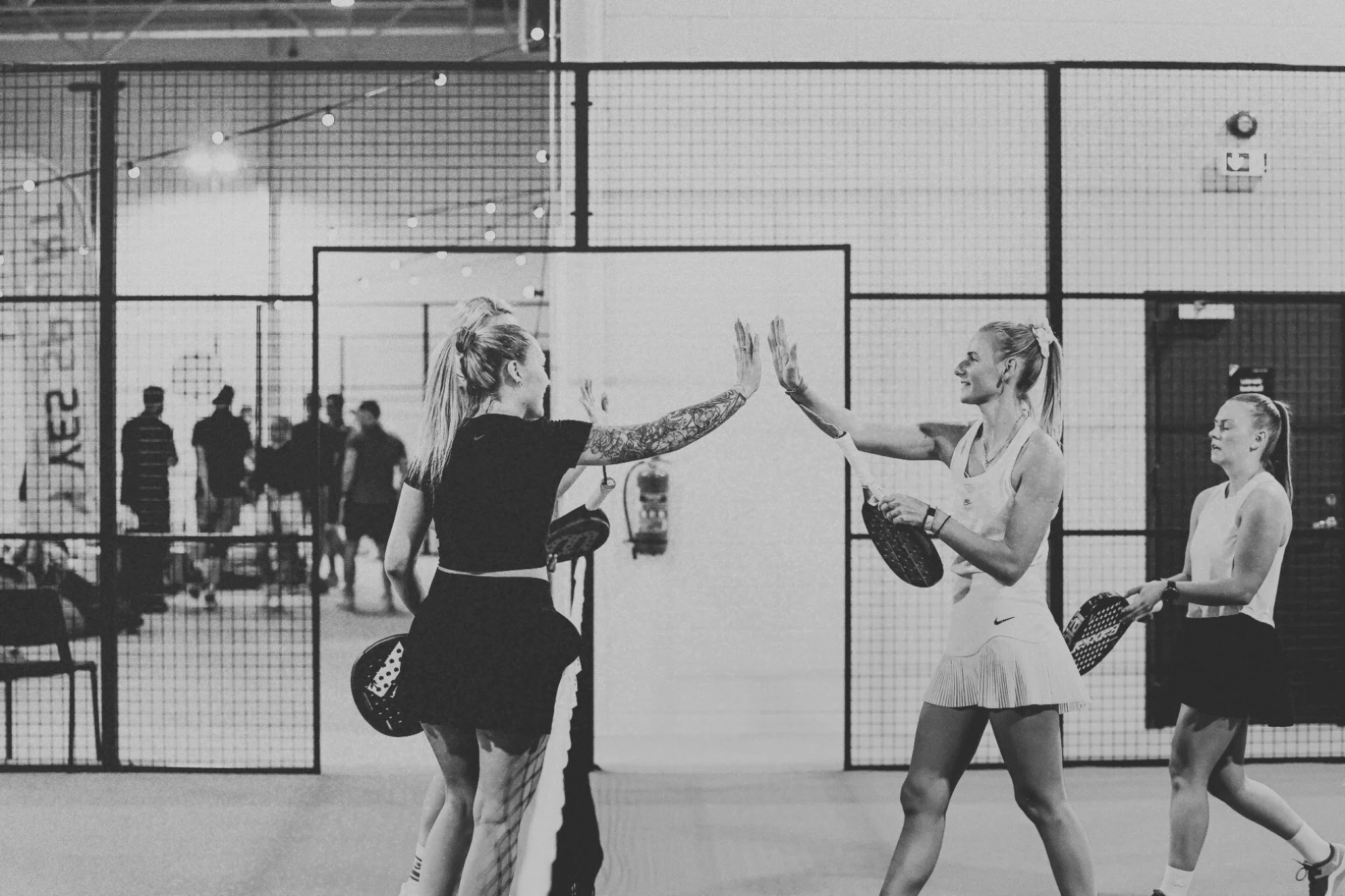 Two women high-five after playing padel on an indoor court, with two other players in the background holding rackets, and a net dividing the court.