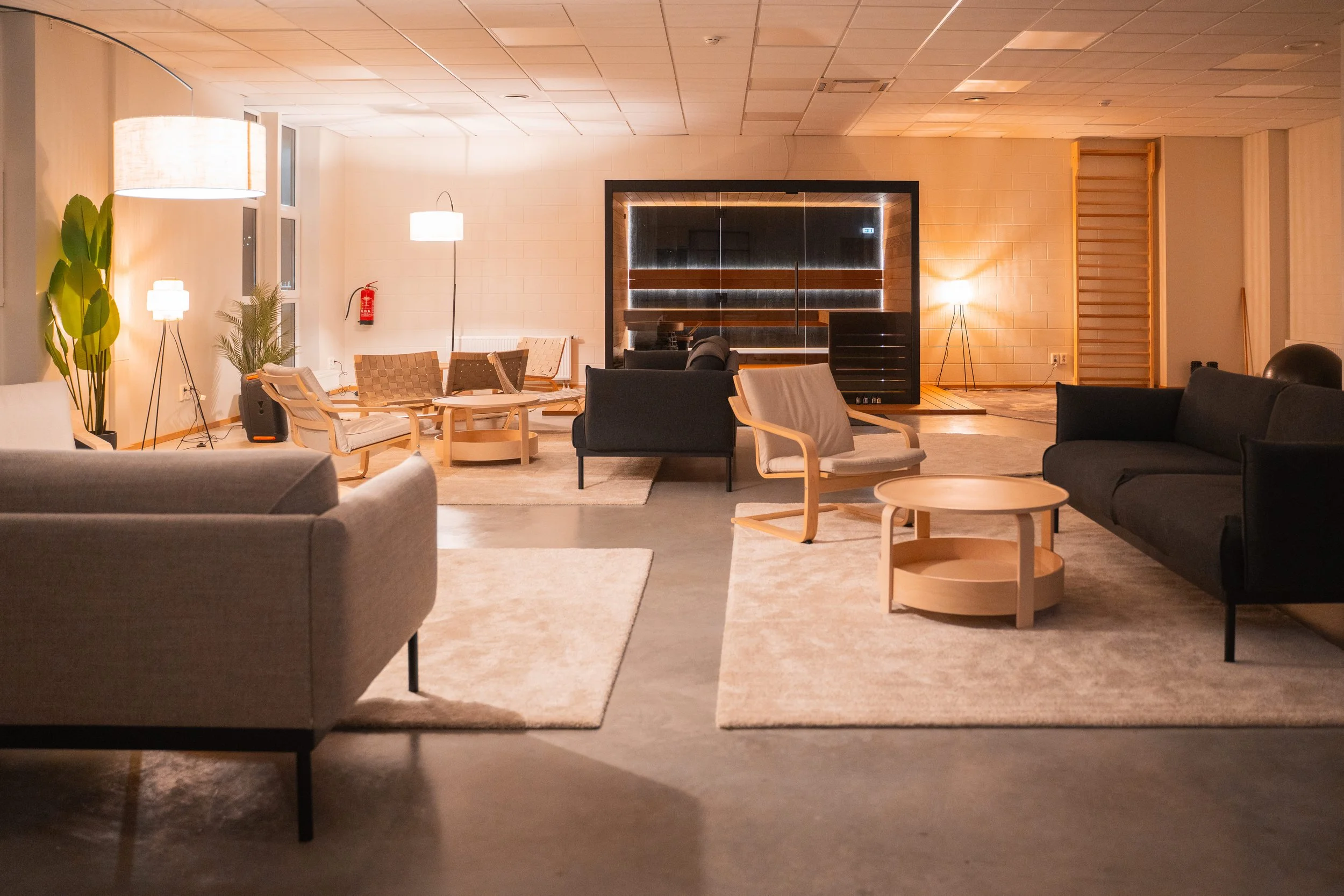 A spacious, modern room with minimalist furniture, including sofas, armchairs, and coffee tables, illuminated by floor and table lamps, and featuring a large mirror and some indoor plants.