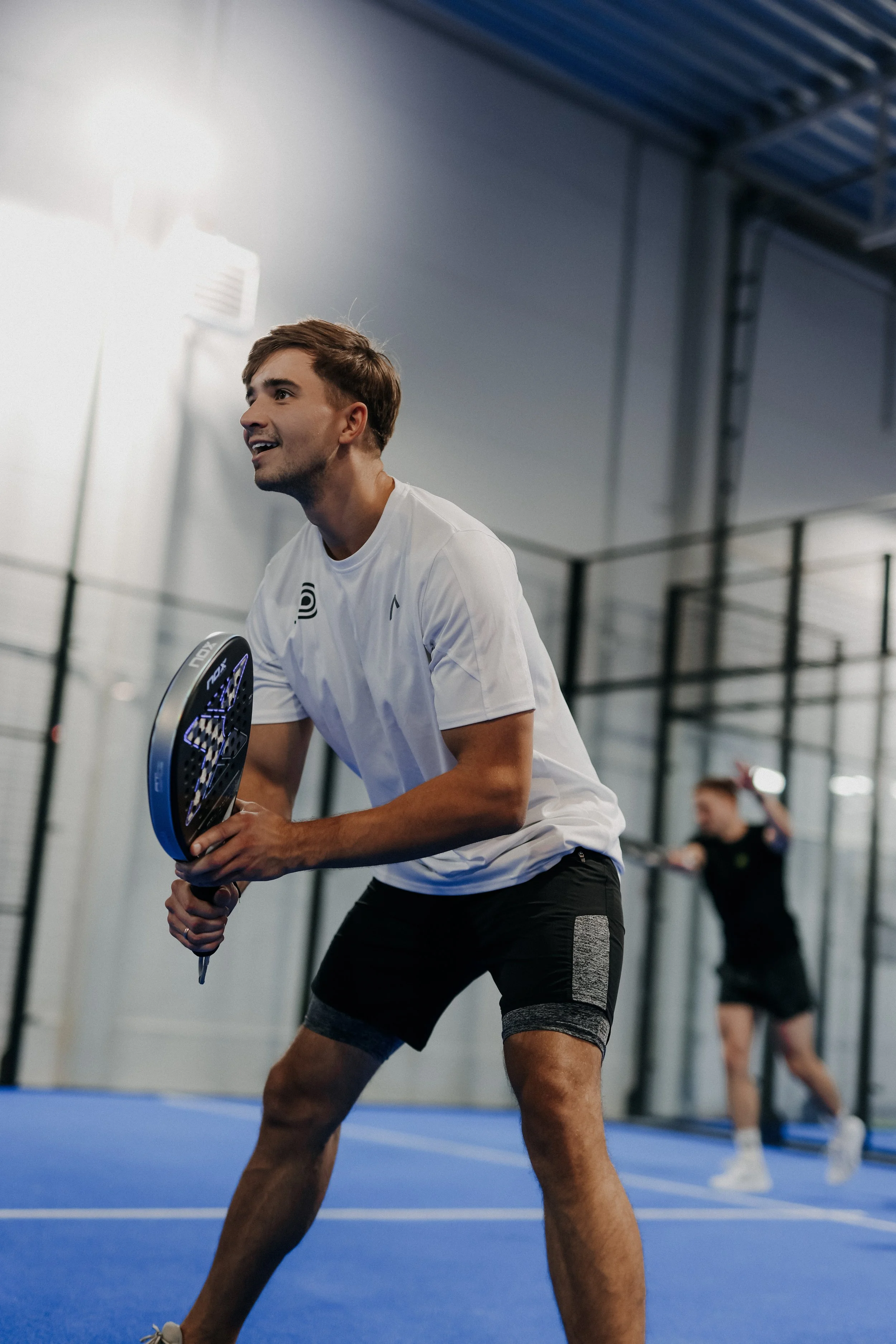 A young man playing padel indoors, holding a racket, with another person in the background preparing to hit a ball.