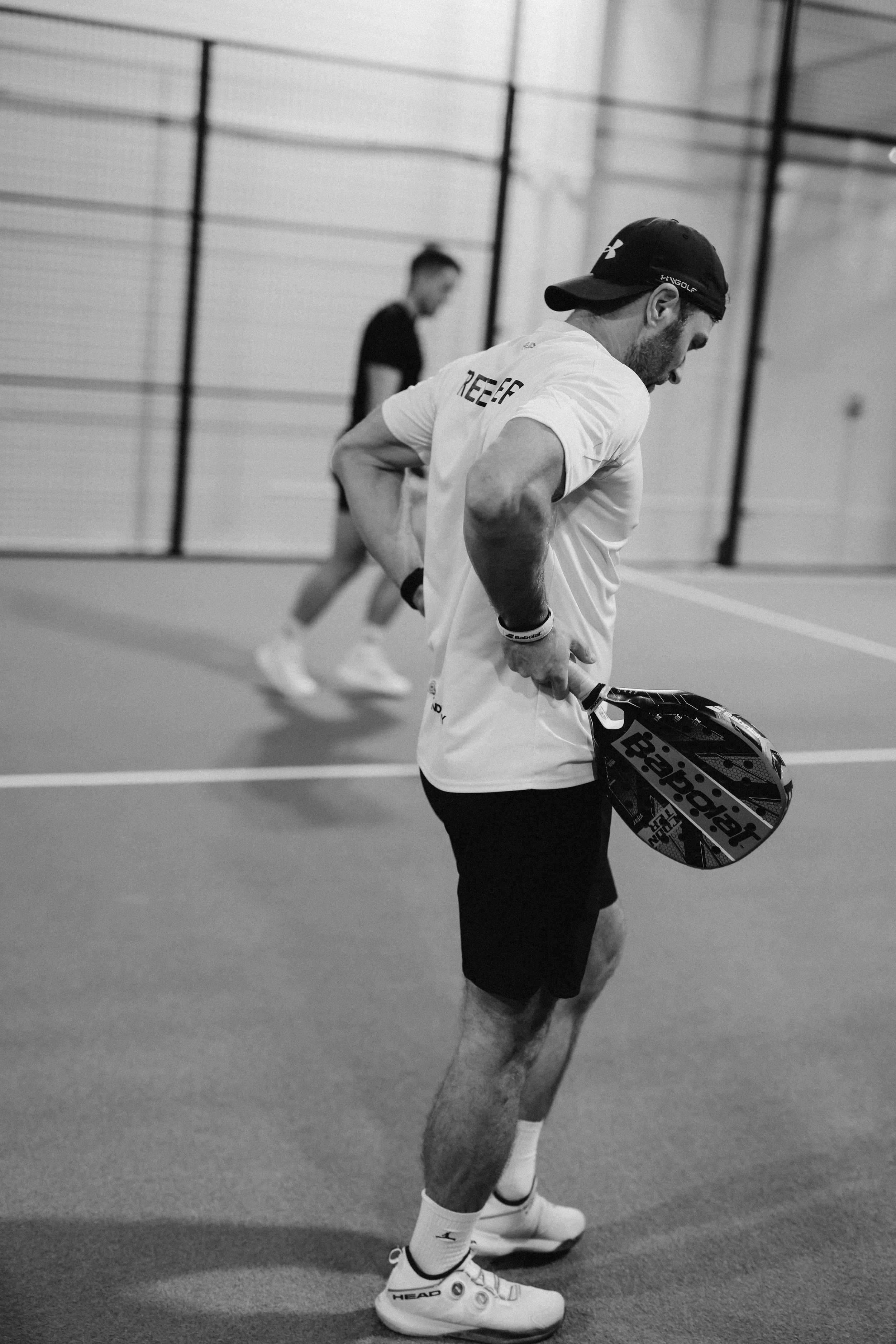 A man in sportswear holding a racket, standing on padel court, with another person in the background practicing.