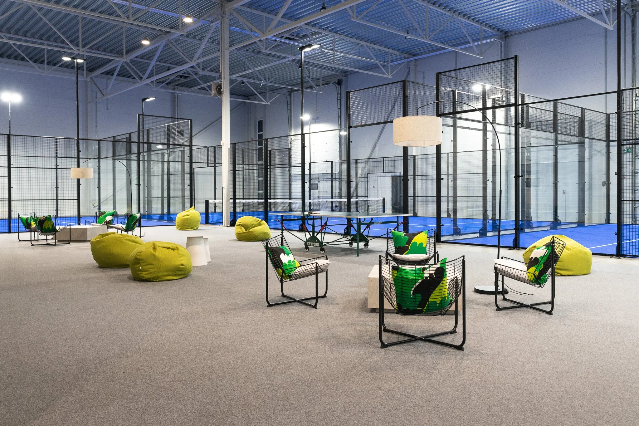 Indoor sports facility with multiple court sections for padel, surrounded by black metal fencing. The space is well-lit with ceiling lights and features seating areas with green and black chairs, bean bags, and decorative pillows with green and black