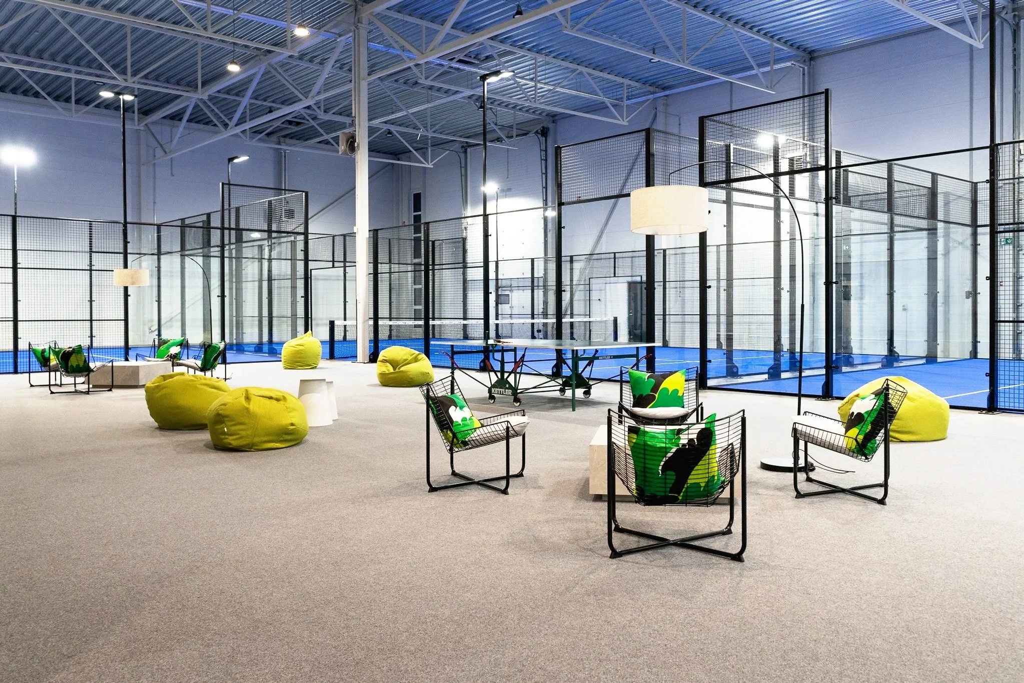 Indoor sports facility with a ping pong table, bean bag chairs, lounge chairs with fondant pillows, and a padel court with glass walls.