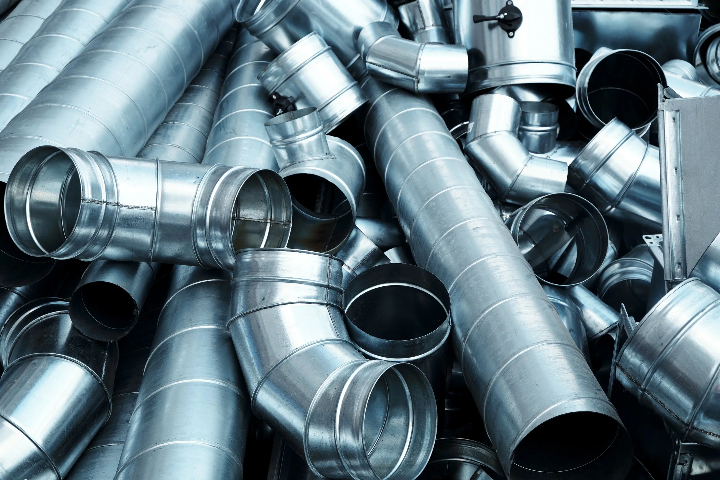 A pile of metal ventilation ducts and pipes, some with fittings and elbows, stacked in a warehouse or workshop.