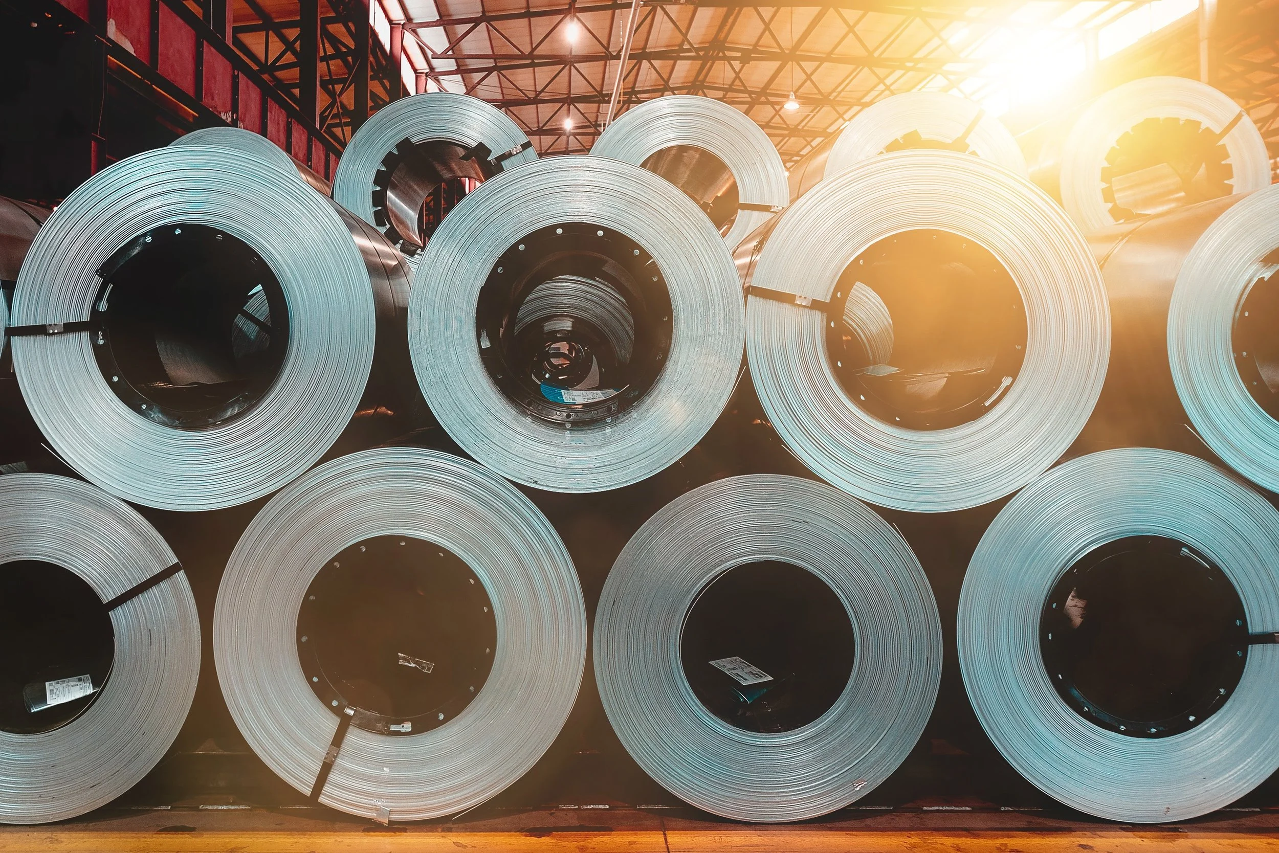 Multiple large steel coils stacked in a warehouse with sunlight streaming in from the side.