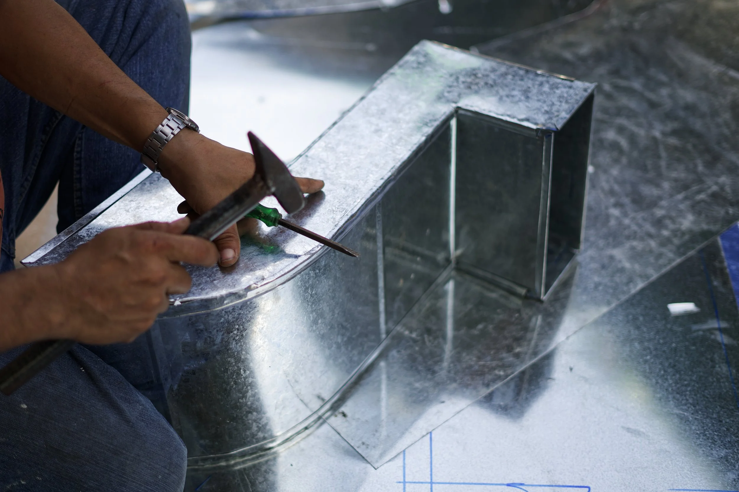 Someone is working on a metal project, using a hammer and screwdriver on a large sheet of metal and a metal box.