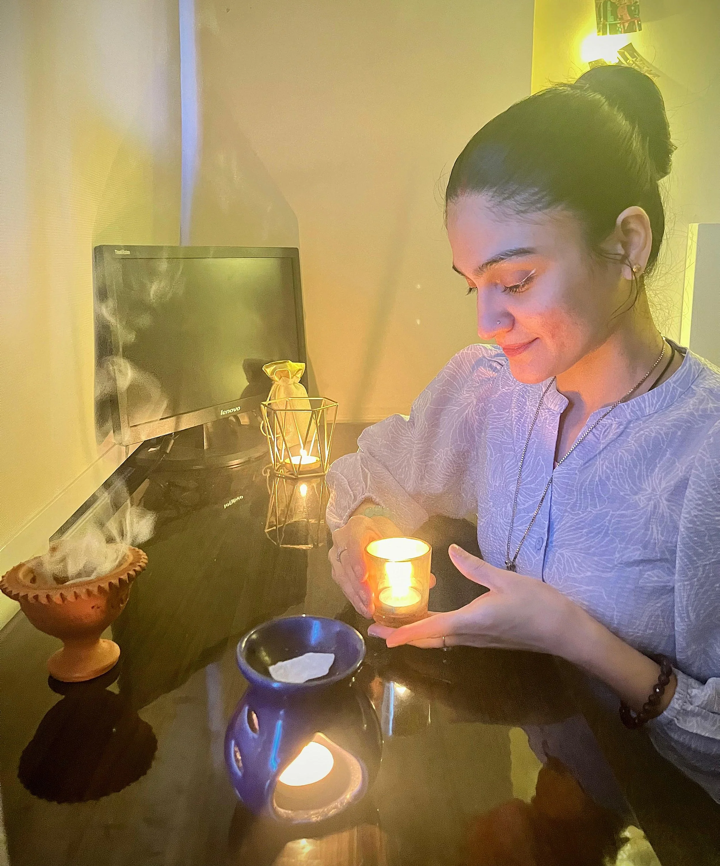 A woman with dark hair in a bun, smiling softly, holding a lit candle in a glass holder, sitting at a dark wooden table with a burning incense holder and a small blue ceramic pot. The background features a monitor, candle, and yellow lighting.