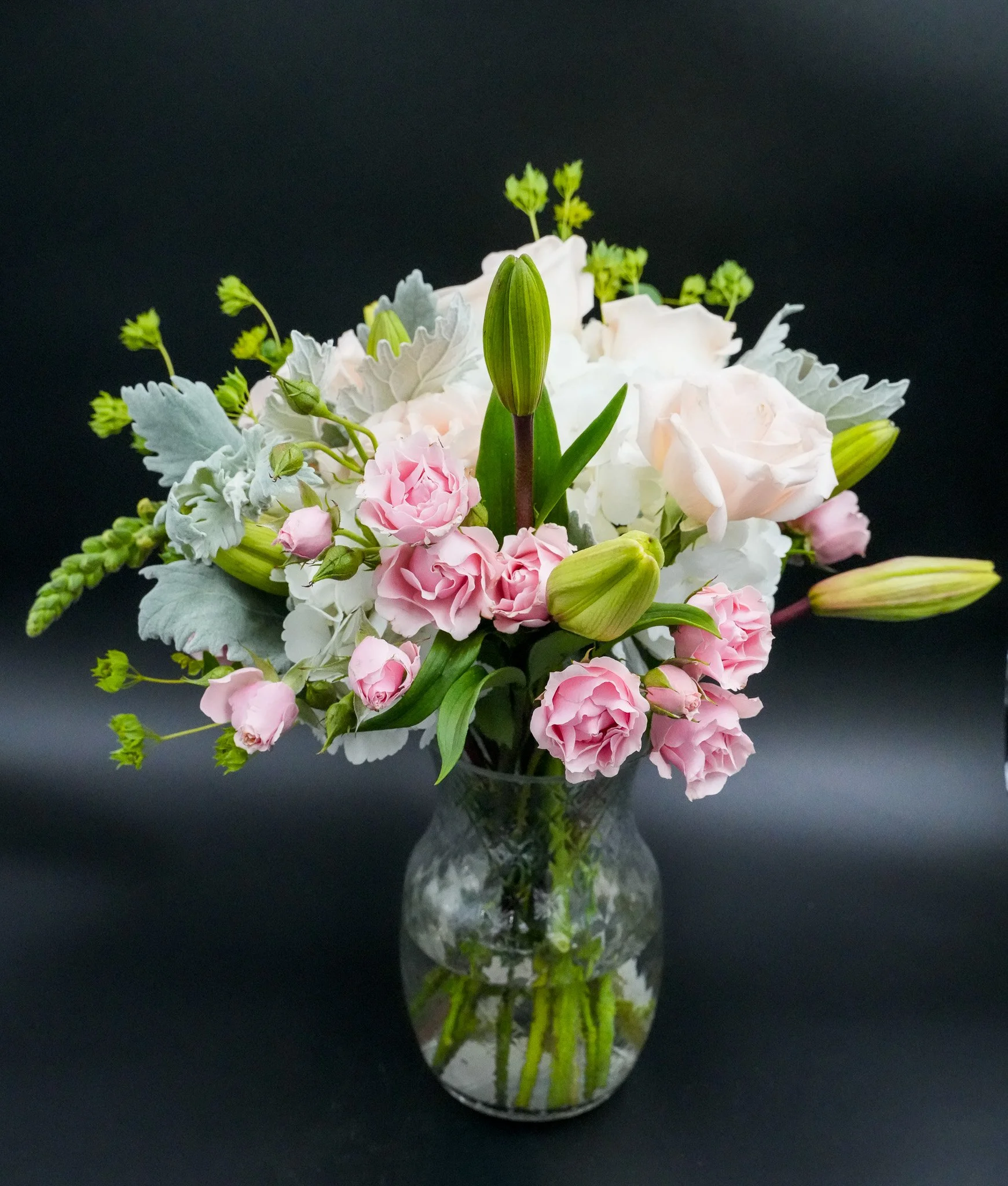A glass vase filled with a bouquet of pink, white, and green flowers, including lilies, carnations, and greenery, set against a black background.