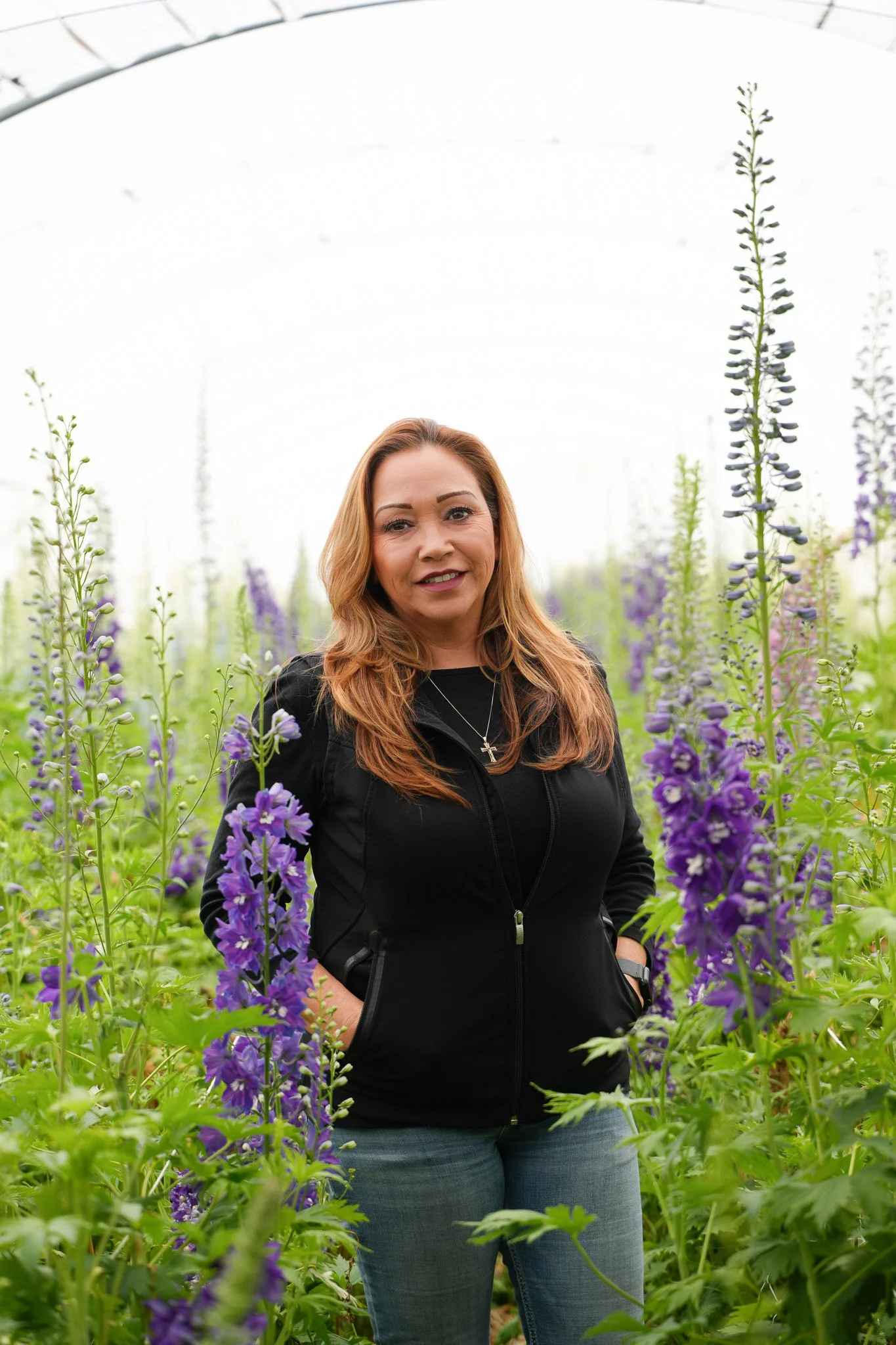 A woman with long reddish-brown hair wearing a black jacket and jeans, standing in a field of tall purple flowers, smiling at the camera.