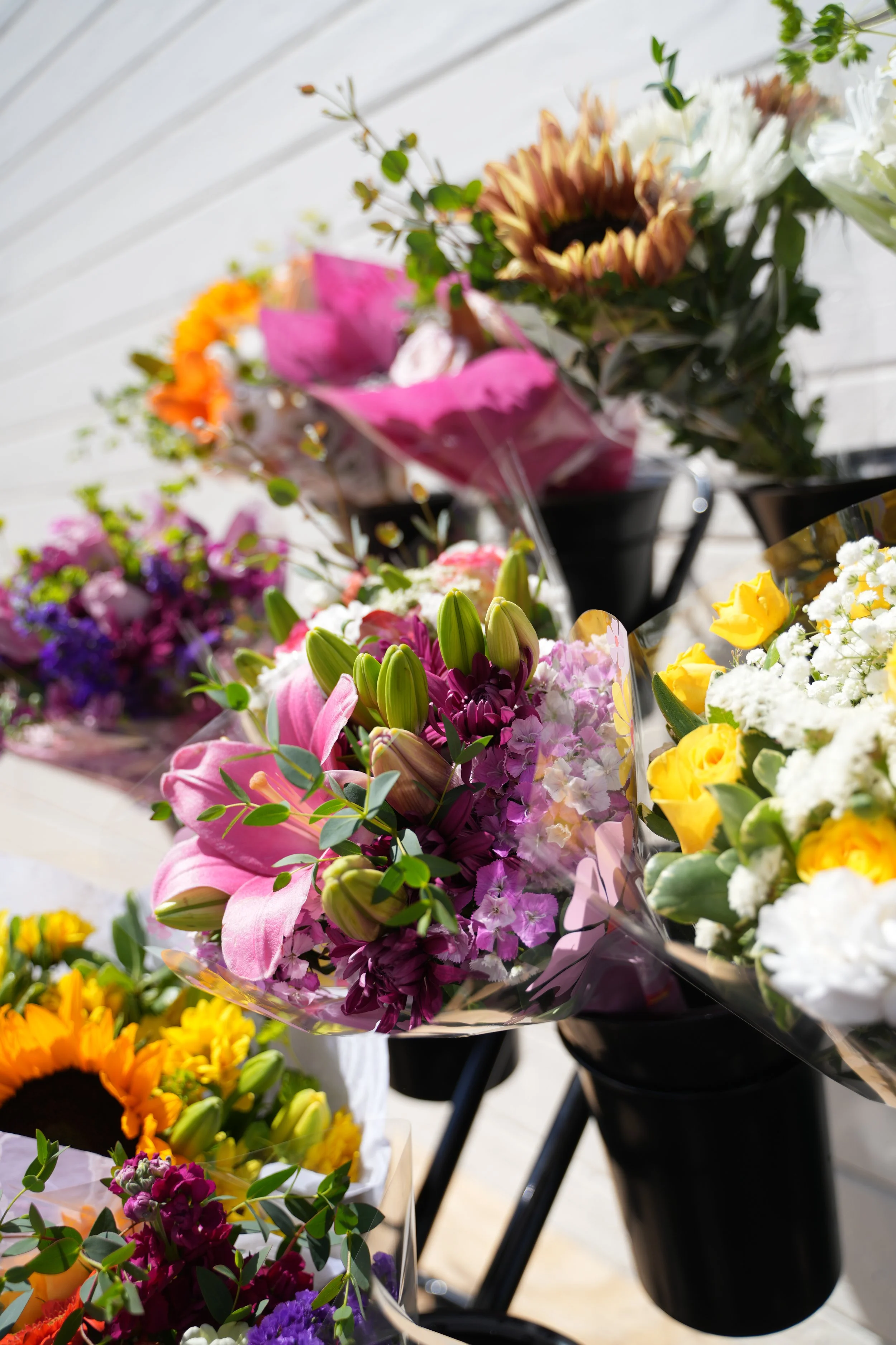 Display of various colorful flower bouquets, including pink lilies, purple and white carnations, yellow roses, and sunflowers, arranged in black and clear wrapping, set against a white wooden background.