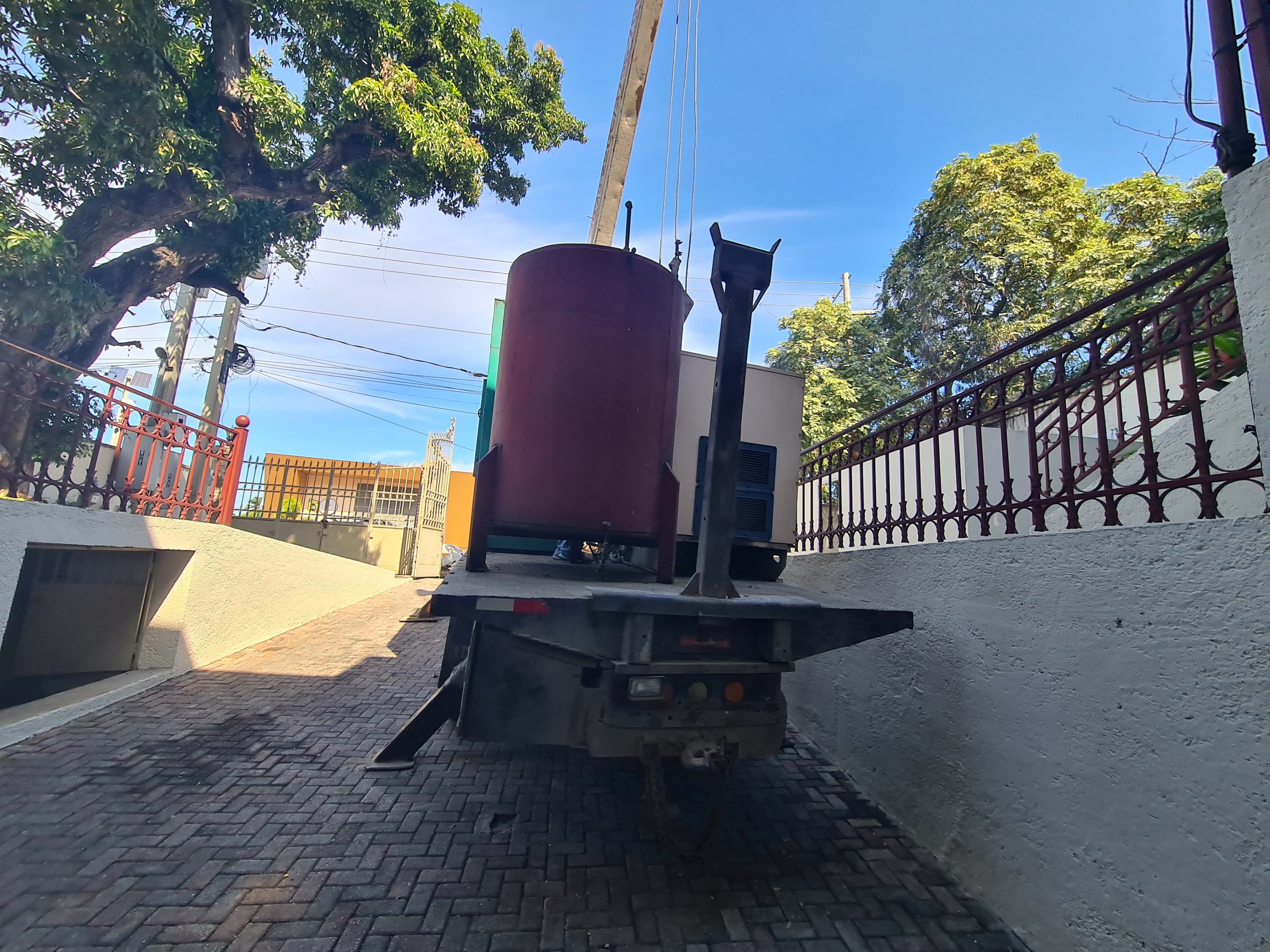 A utility truck with equipment on the back, parked in a driveway with brick paving, bordered by white and red fences, under a blue sky with trees.