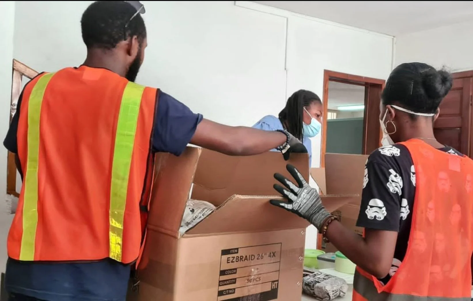 Three people wearing safety vests and masks inspecting a large cardboard box indoors, with one person holding the box open for another person to look inside.