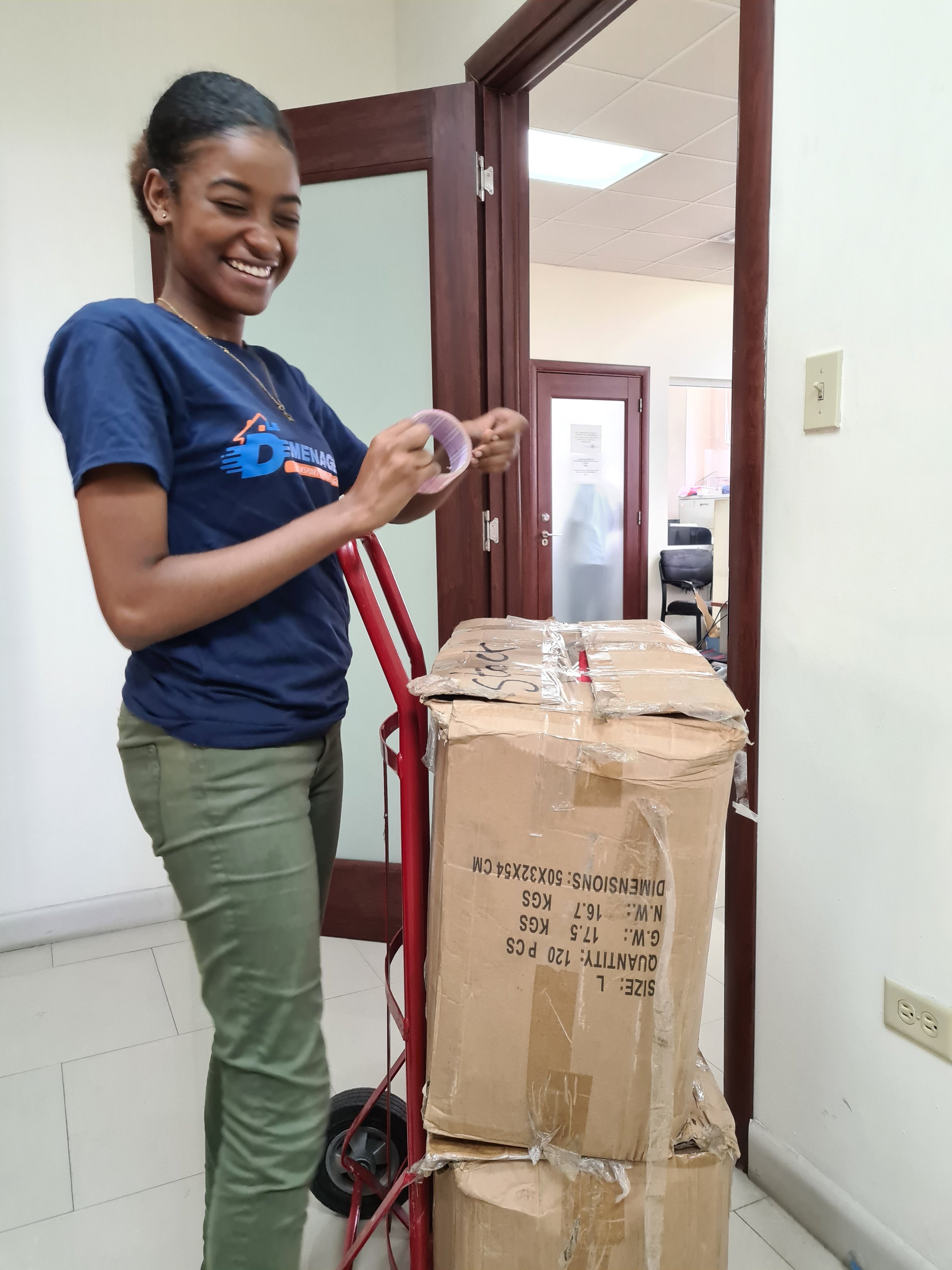 A young woman smiling while handling packing tape next to a red hand truck with boxes in an office setting.