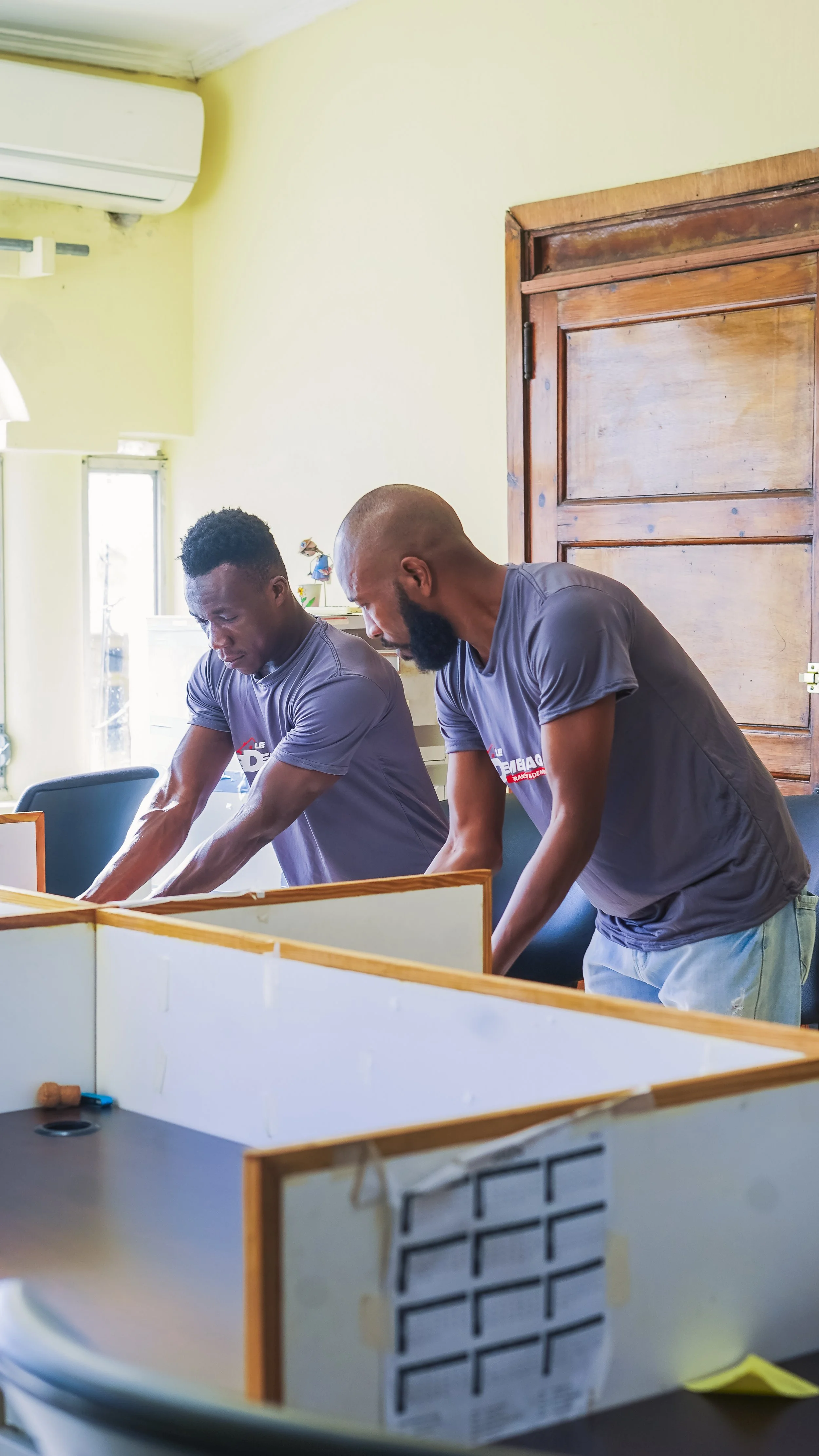 Two men working together at a desk in a room with light green walls and a wooden door, both wearing matching gray T-shirts.