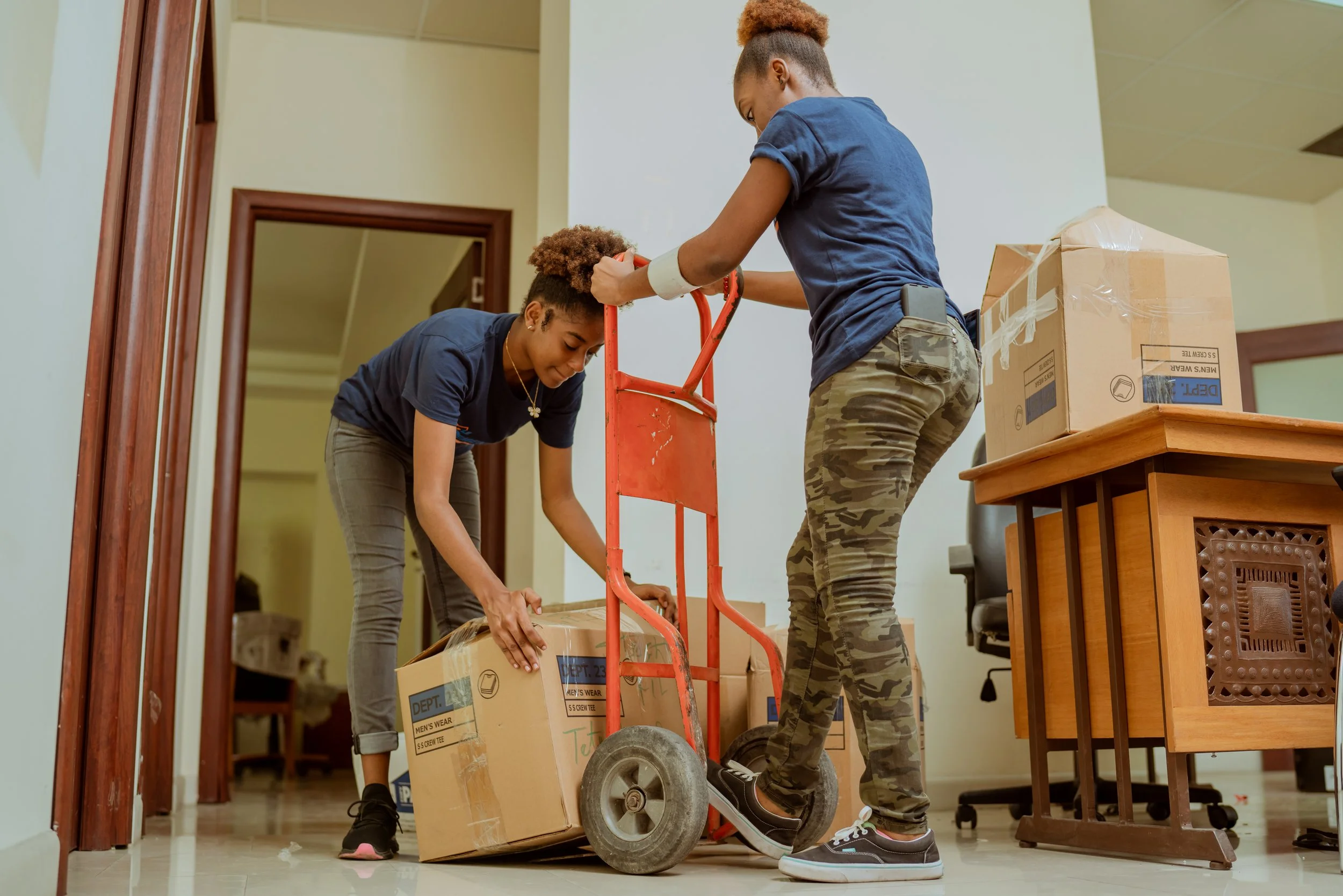 Two women moving boxes using a hand truck in an indoor space.