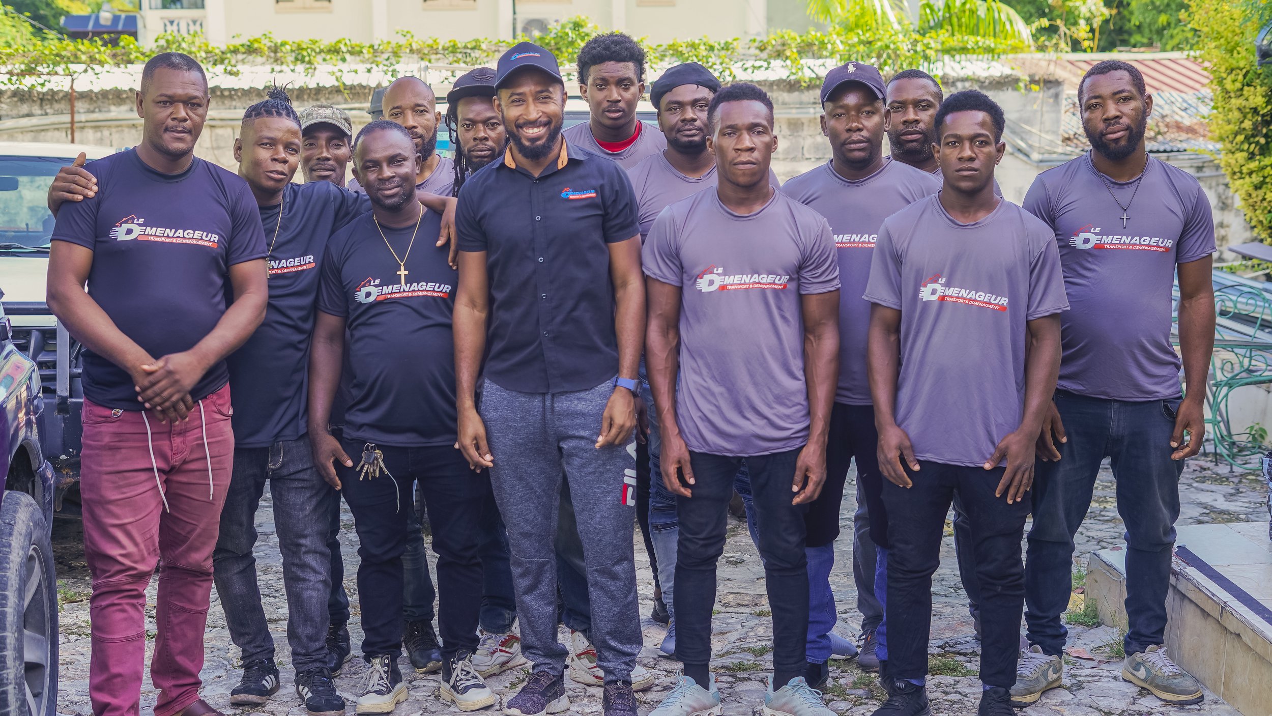 Group of men standing together outdoors, some wearing gray T-shirts with a logo, two wearing black shirts, posing for a photo, with greenery and buildings in the background.