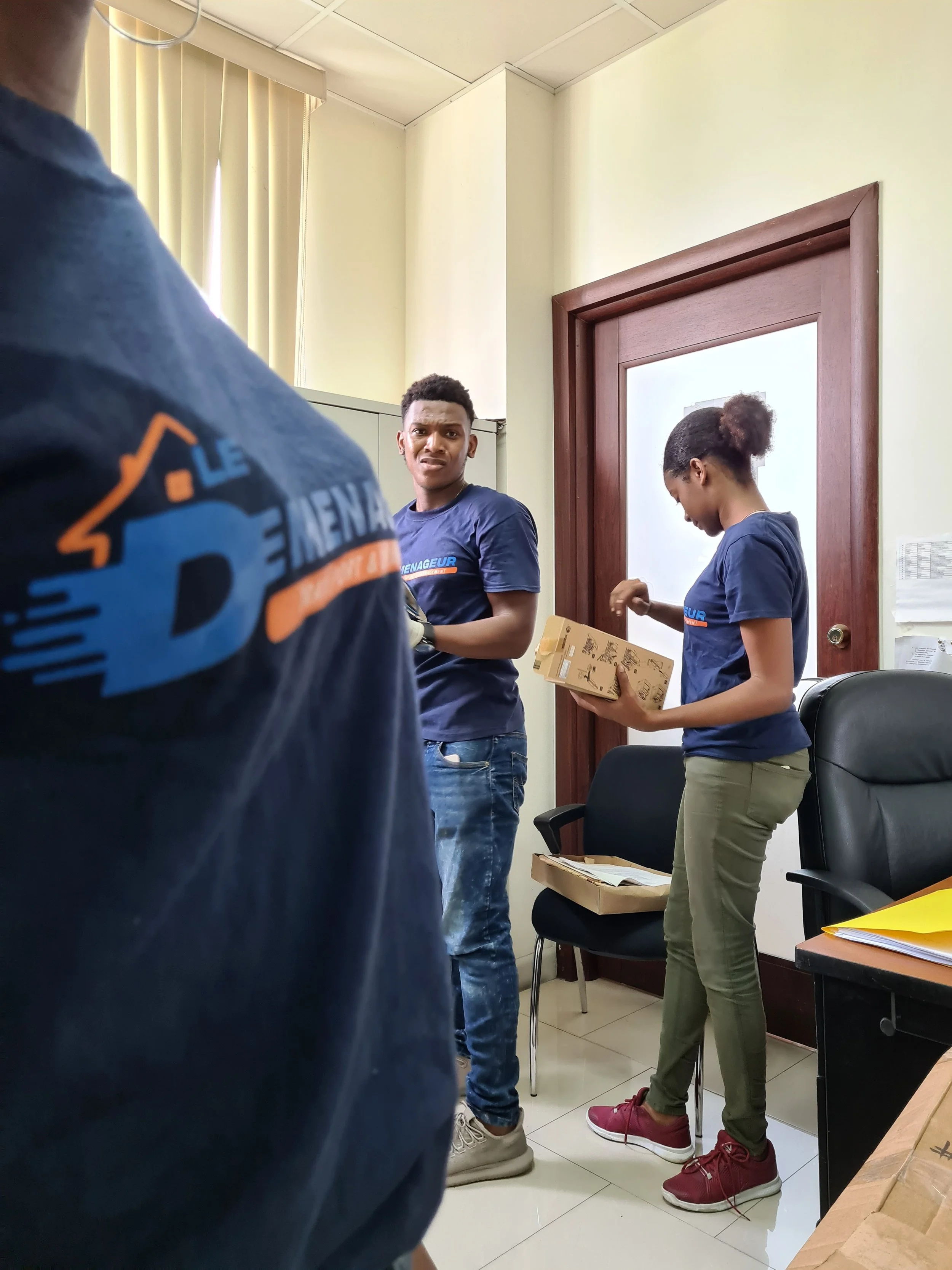 Two people wearing matching navy blue shirts in an office, one man looking confused or surprised and a woman reading a box.