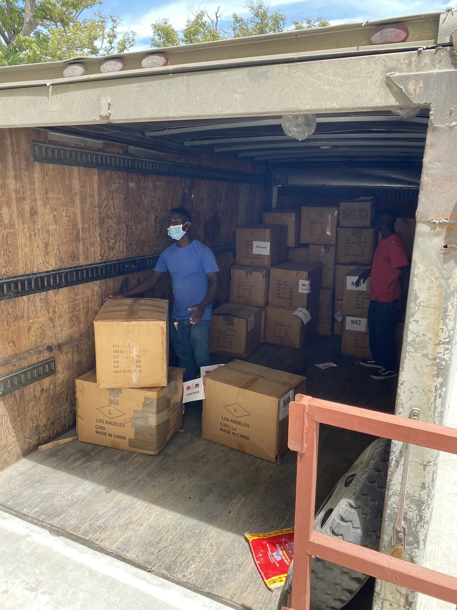 Two men wearing face masks loading or unloading cardboard boxes inside a truck with wooden interior walls.