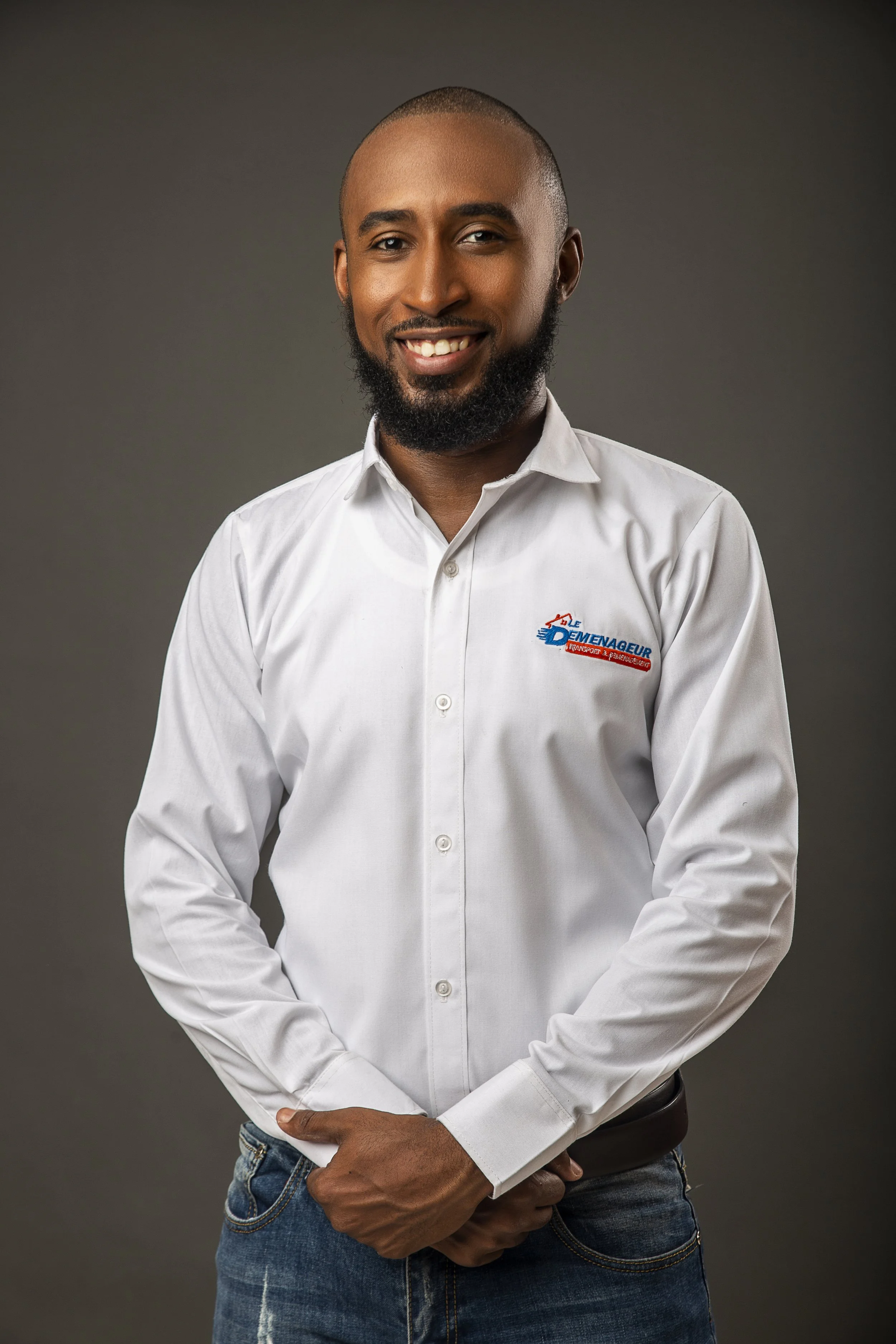A man with a beard and short hair smiling, wearing a white button-up shirt with a logo and blue jeans, standing against a dark background.