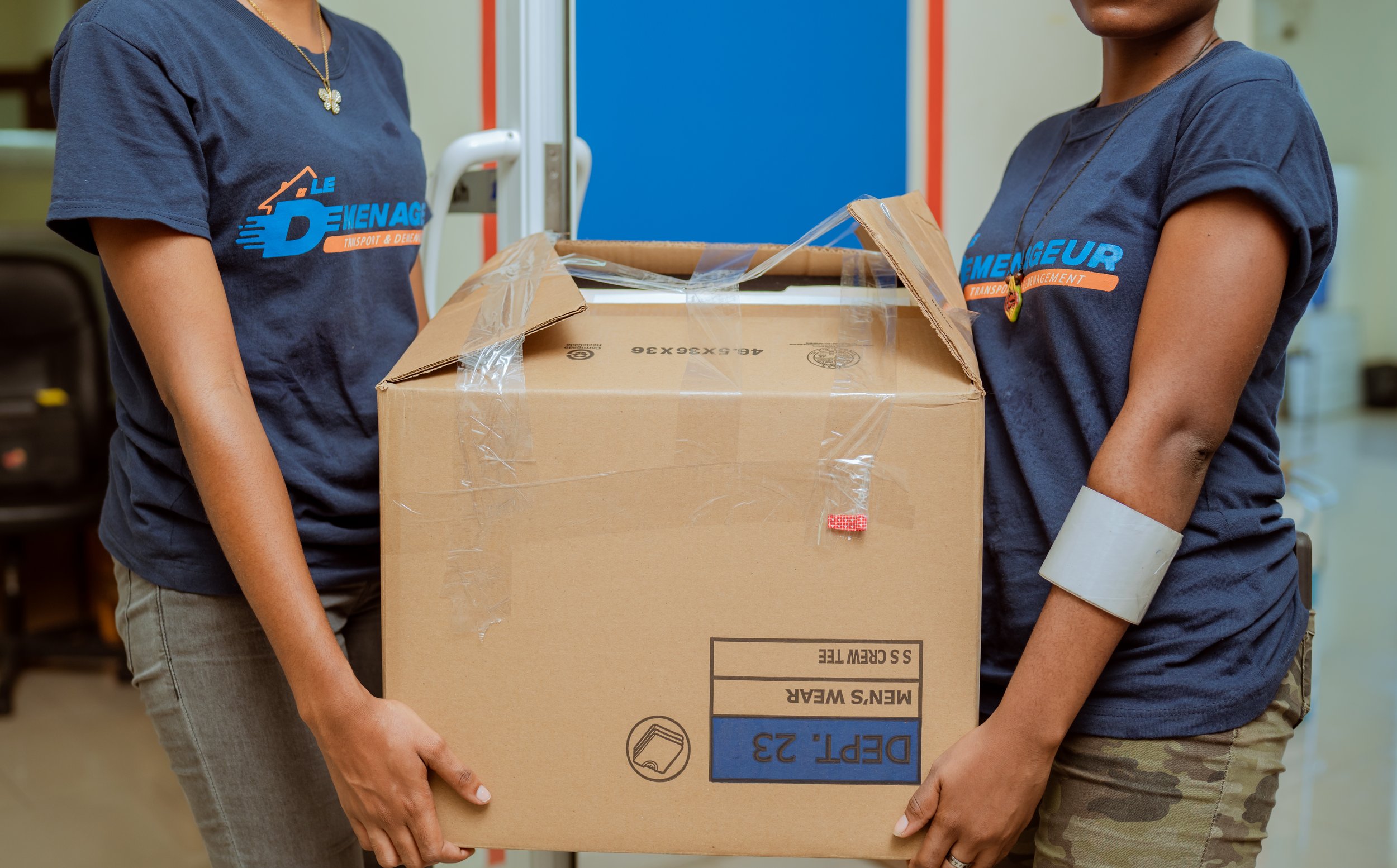 Two people in navy blue t-shirts lifting a large cardboard box.