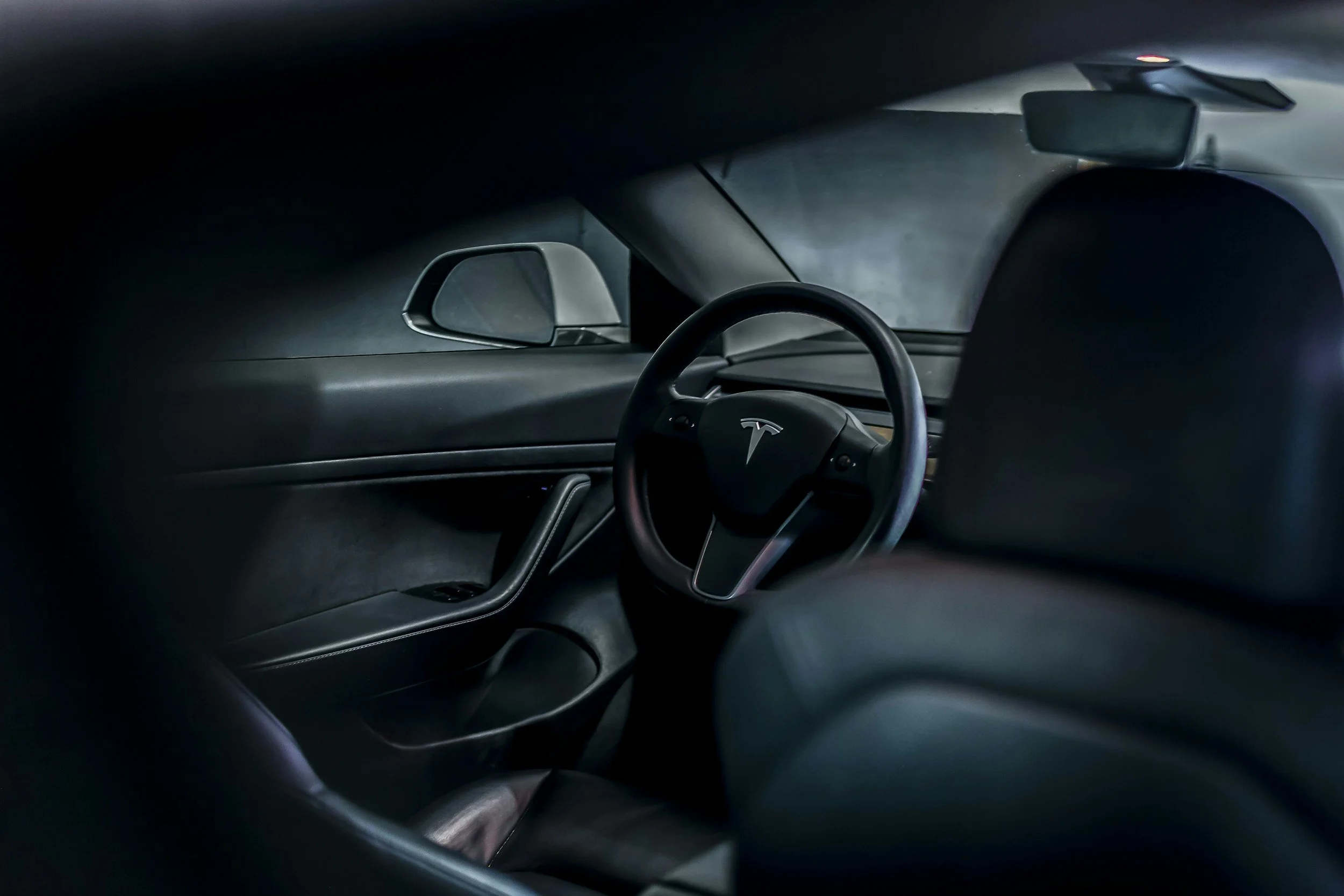 Interior of a Tesla electric car, showing the steering wheel with Tesla logo, black dashboard, and side mirror.