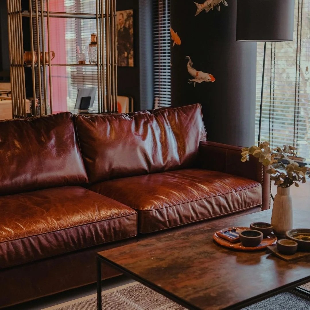 A living room with a brown leather sofa, a wooden coffee table with a vase of dried flowers, and a decorative wall with fish art and wooden blinds on the window.