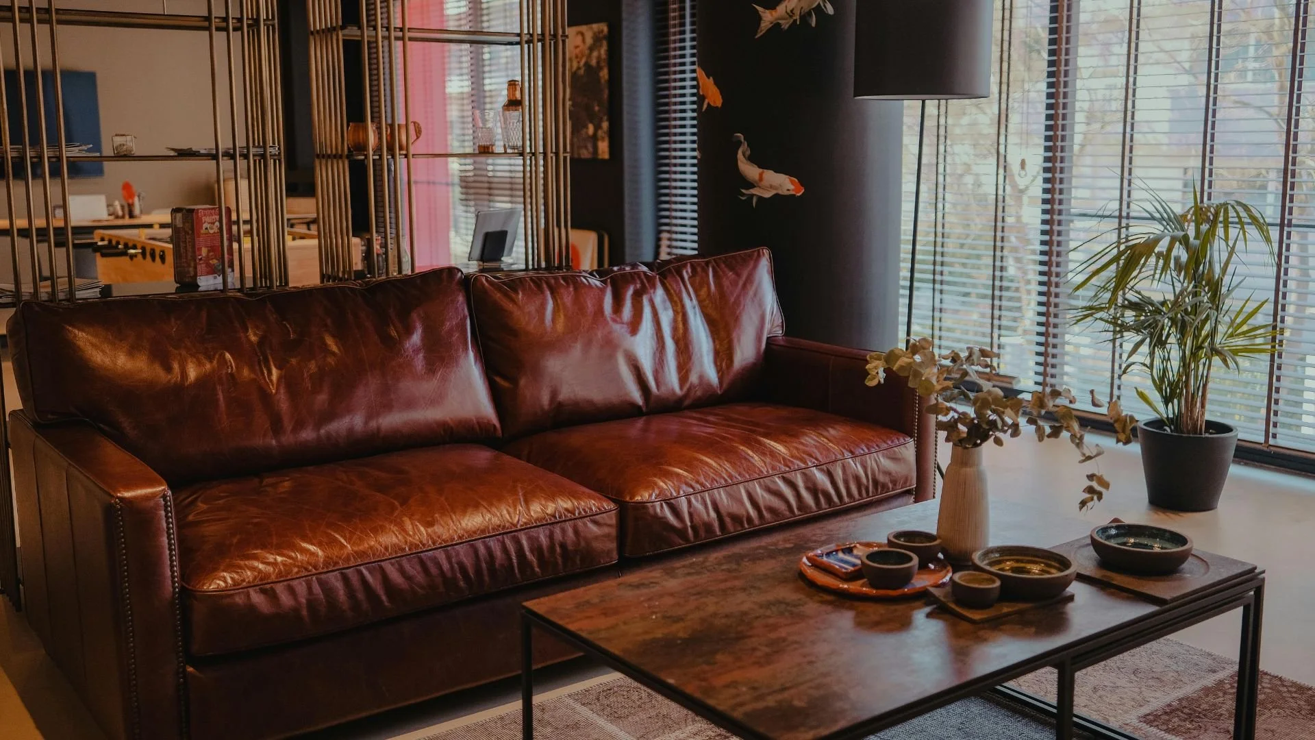 Living room with a brown leather sofa, a wooden coffee table with bowls and a vase, and potted plants near large windows with blinds.