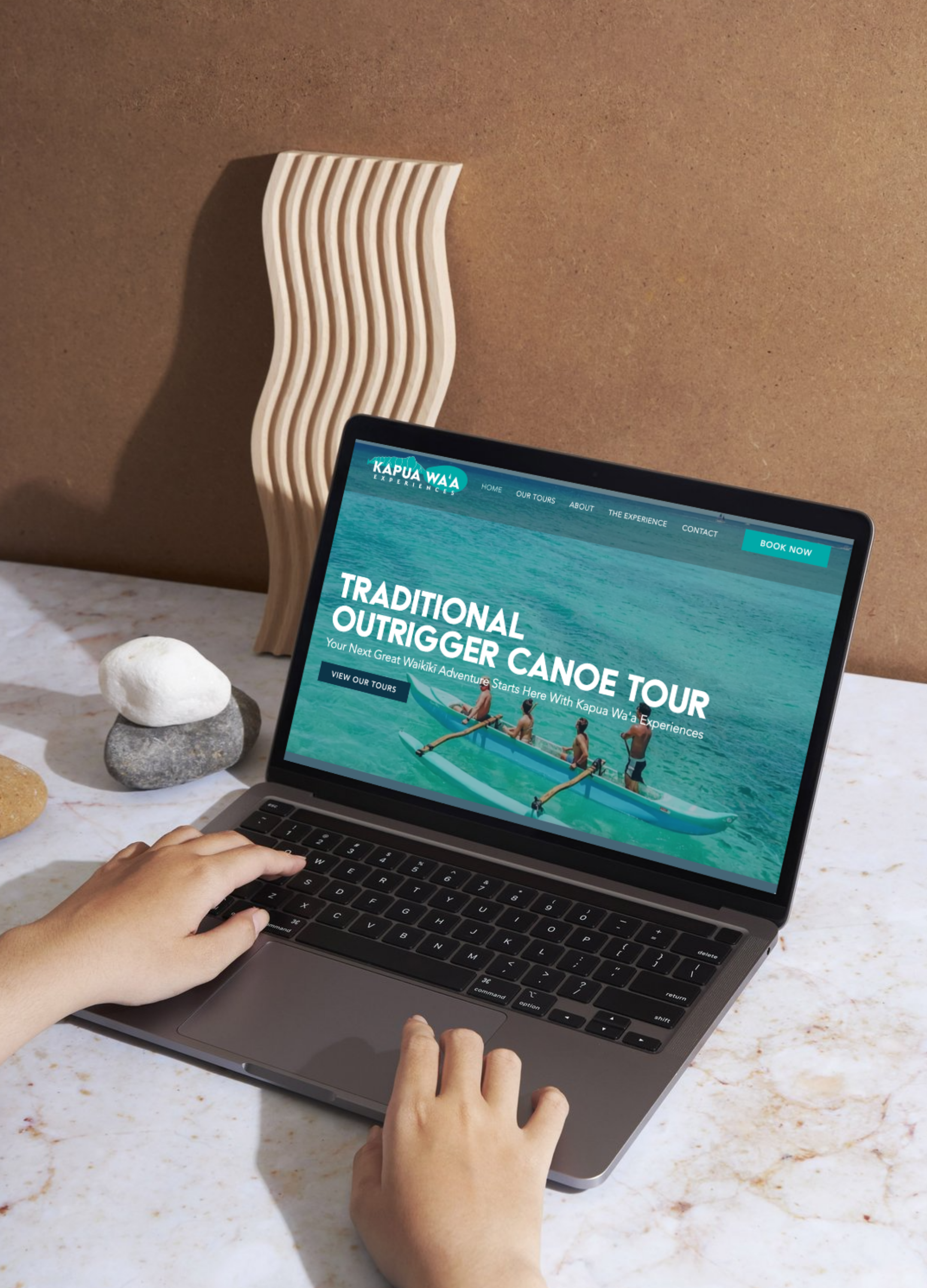 A person’s hands using a laptop on a marble table, displaying a website for Kapua Waa Experiences, promoting a traditional outrigger canoe tour in Waikiki, with decorative stones and a sculptural piece in the background.