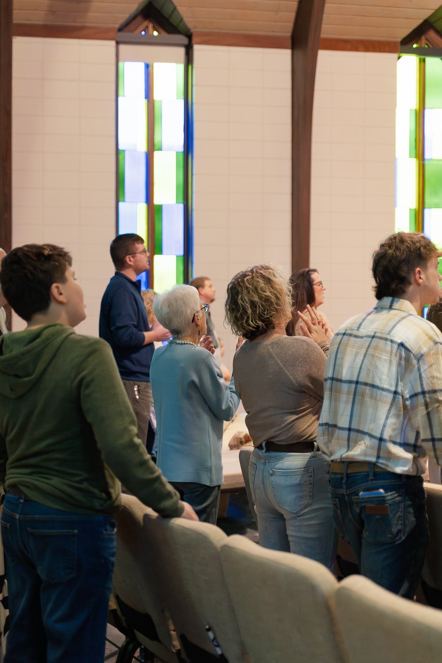 People inside a church or chapel, standing and praying, with stained glass windows in the background.