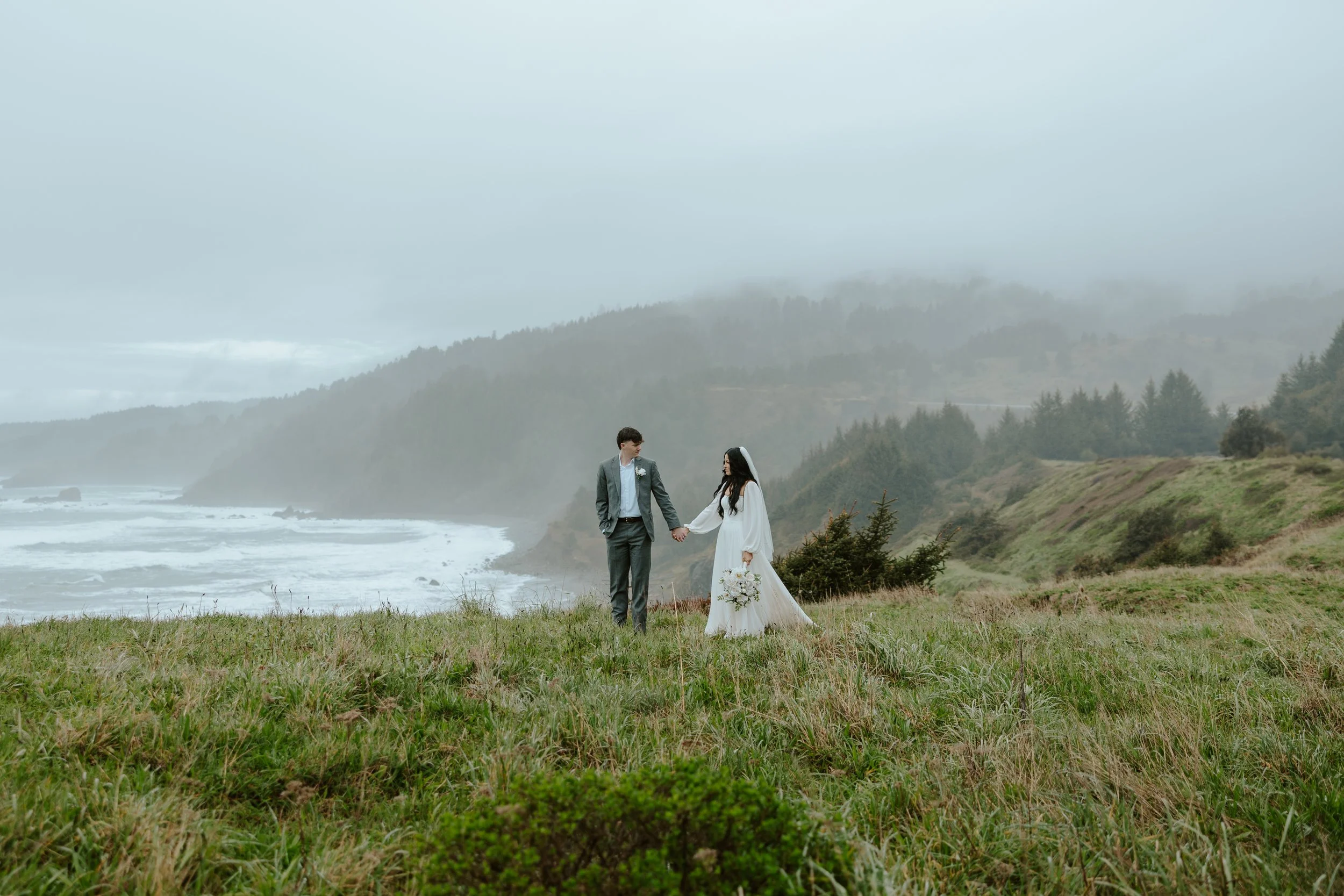 A bride and groom holding hands on a grassy cliff by the ocean with misty hills in the background.
