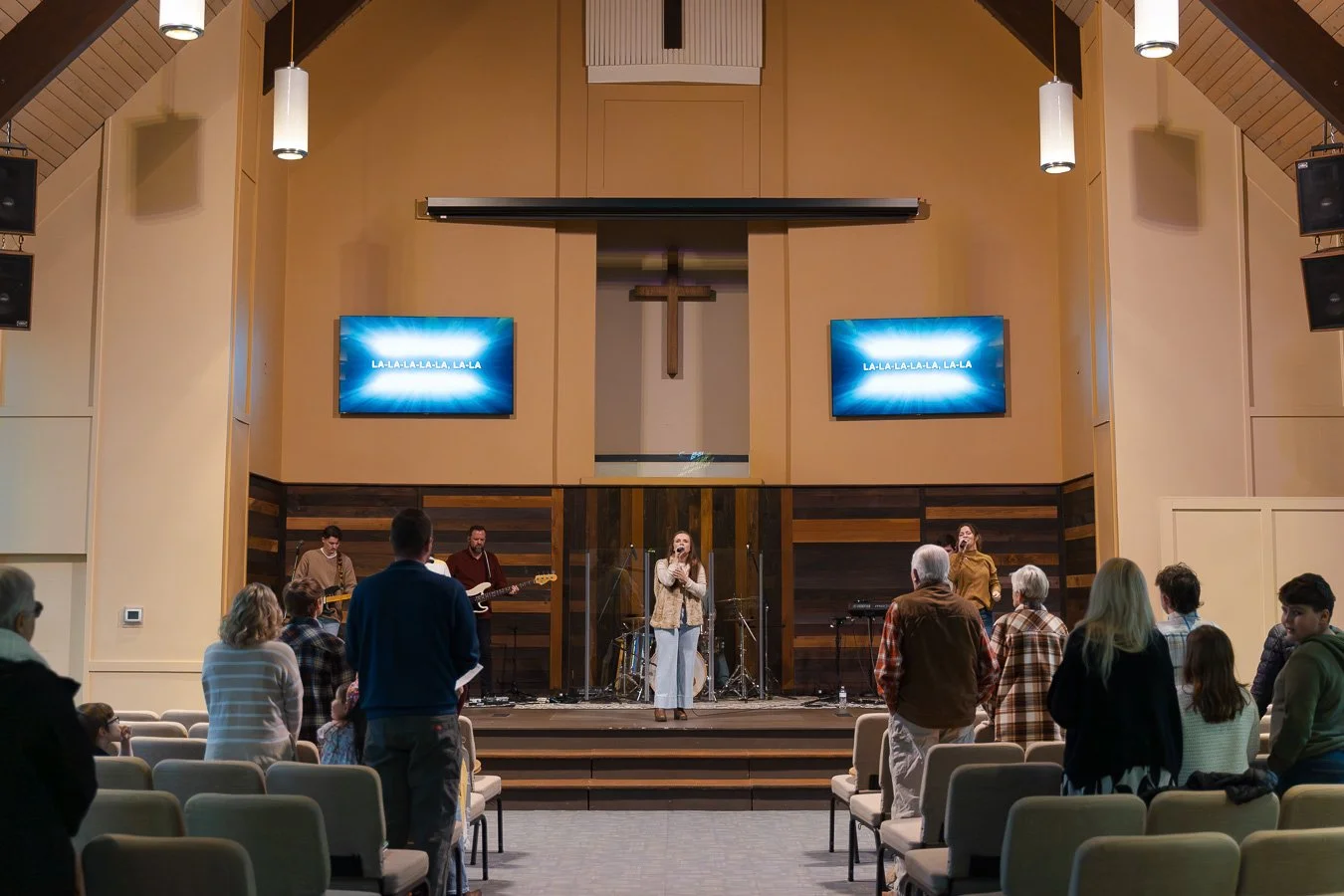 People worshiping inside a church with a band on stage, screens displaying lyrics, and a cross on the wall behind the stage.