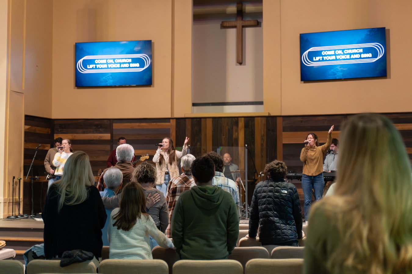 A church congregation gathered for worship, with some standing and singing while a band performs on stage. Two large screens display the message "Come on, Church. Lift your voice and sing." The interior features a cross on the wall behind the stage.