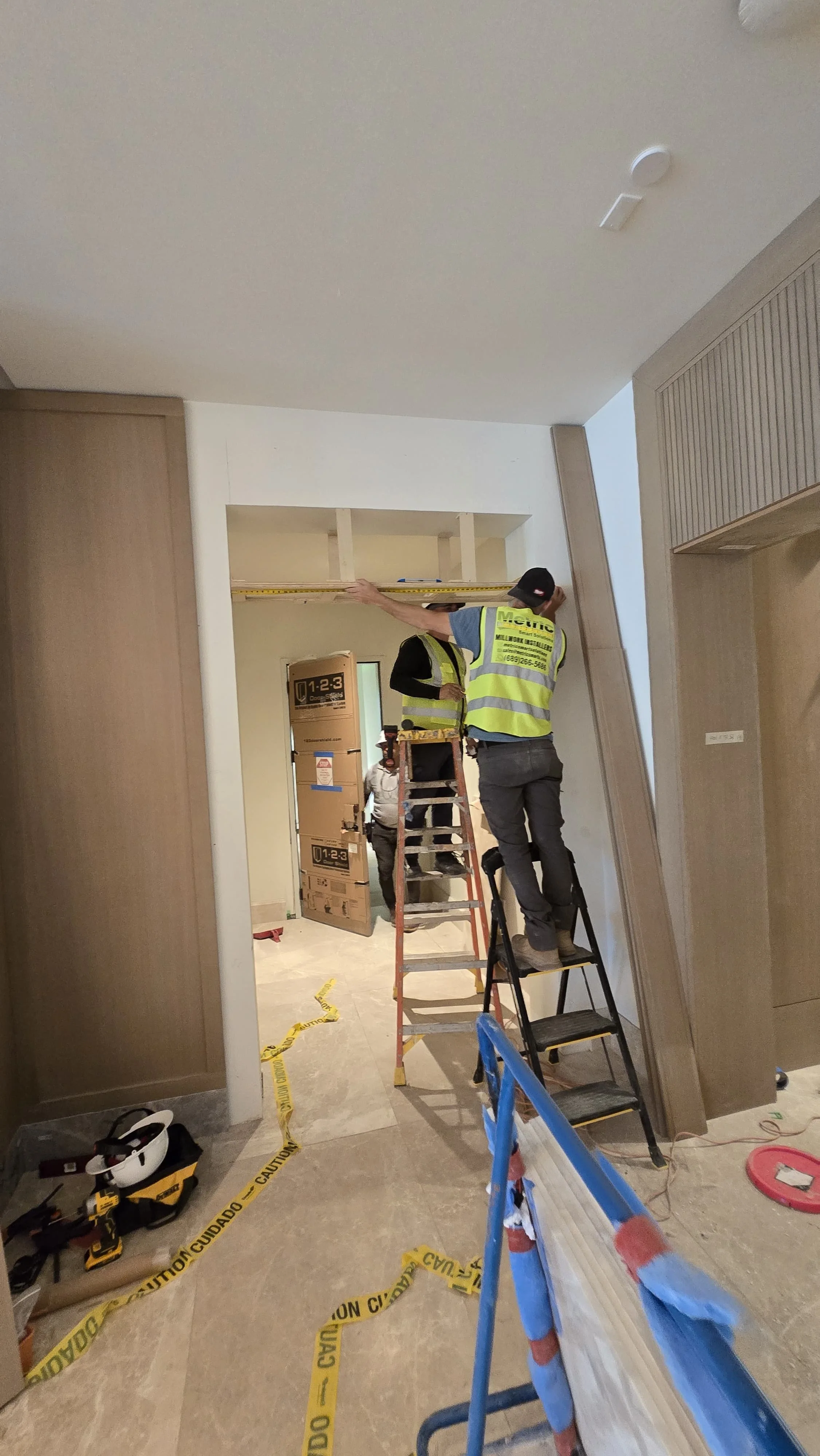 Construction workers installing a ceiling feature inside a building, with caution tape on the floor, tools on the ground, and a ladder in use.