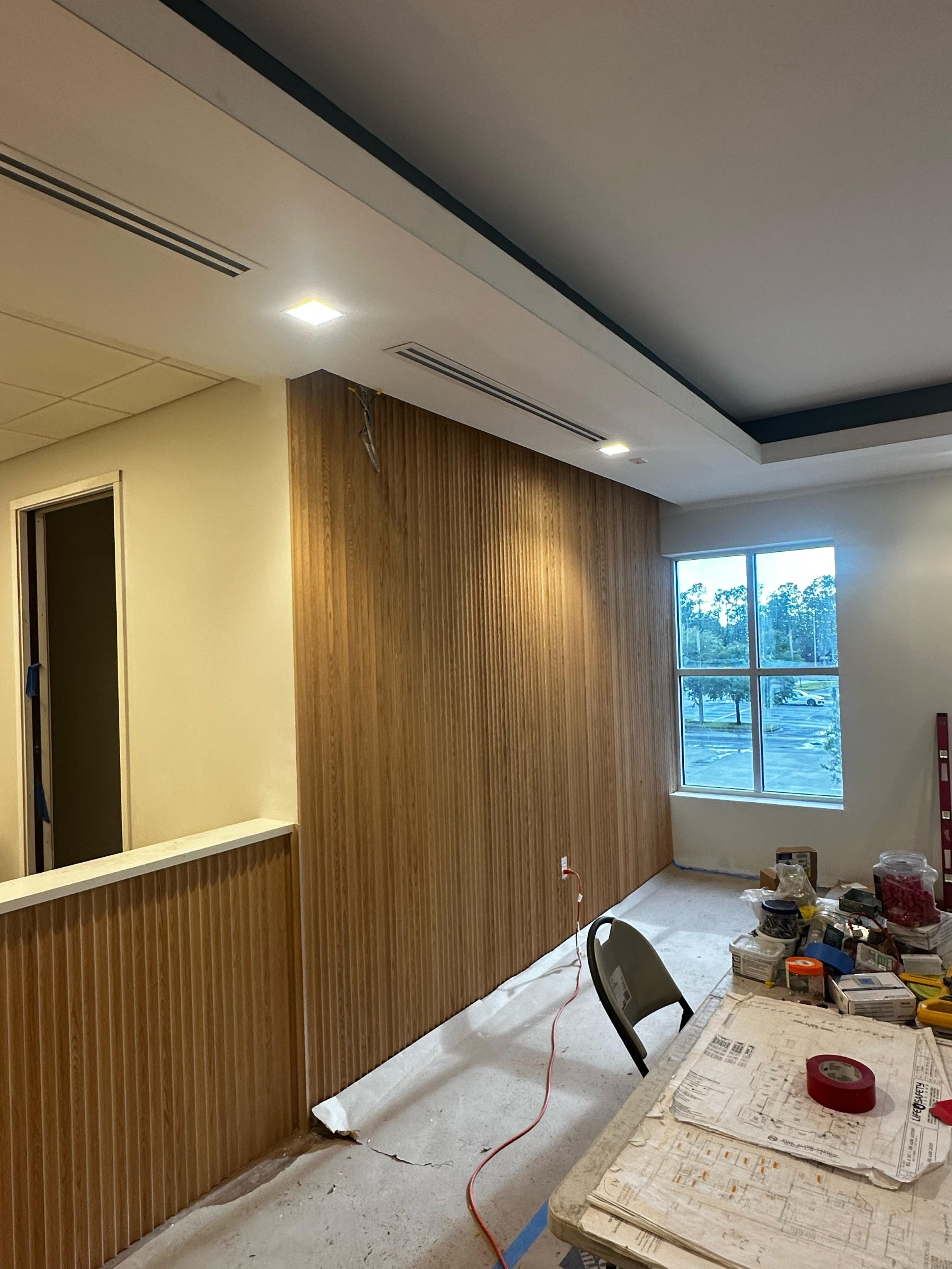 Room under construction with a wood-paneled wall, a window showing an outdoor parking lot with trees, a table with tools and construction materials, and an office chair.